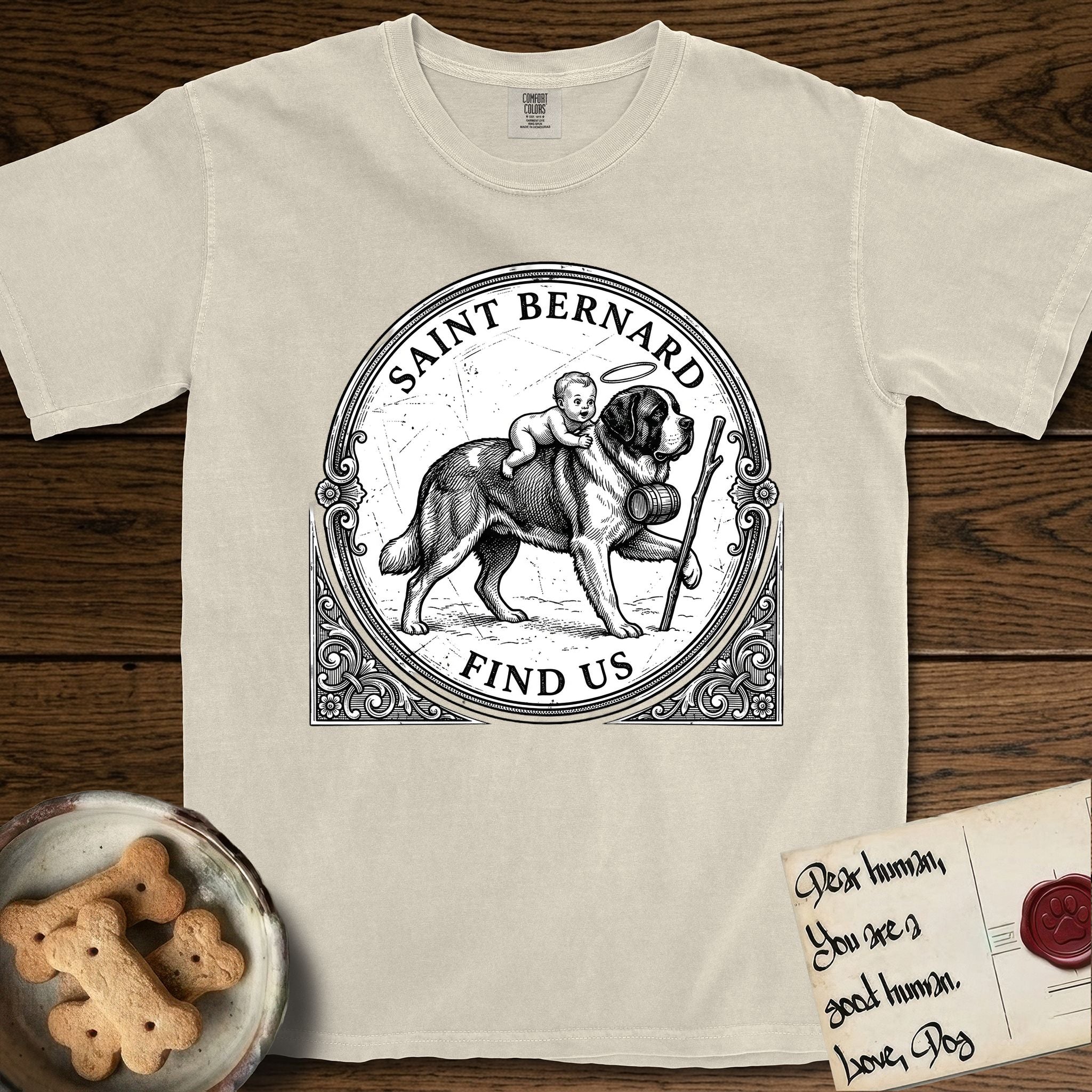 Patron Saint Medallion Saint Bernard T-Shirt flat lay in Ivory, featuring a vintage engraving coin medallion of a haloed Saint Bernard carrying a child across a mountain pass, with the petition FIND US.