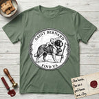 Patron Saint Medallion Saint Bernard T-Shirt flat lay in Military Green, featuring a vintage engraving coin medallion of a haloed Saint Bernard carrying a child across a mountain pass, with the petition FIND US.