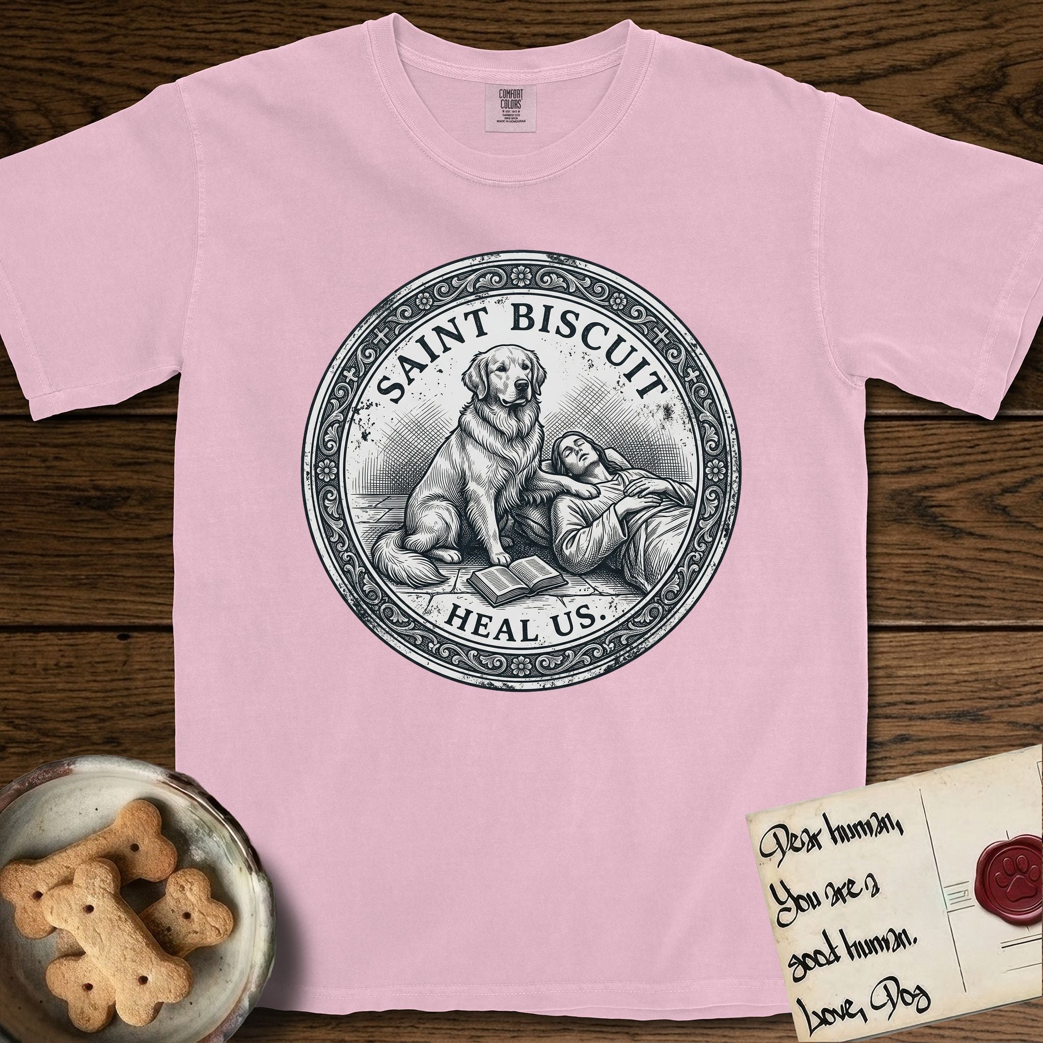 Patron Saint Medallion Saint Biscuit T-Shirt flat lay in Blossom, featuring a vintage engraving coin medallion of a haloed Golden Retriever with a devotee resting against her and an open book, with the petition HEAL US.