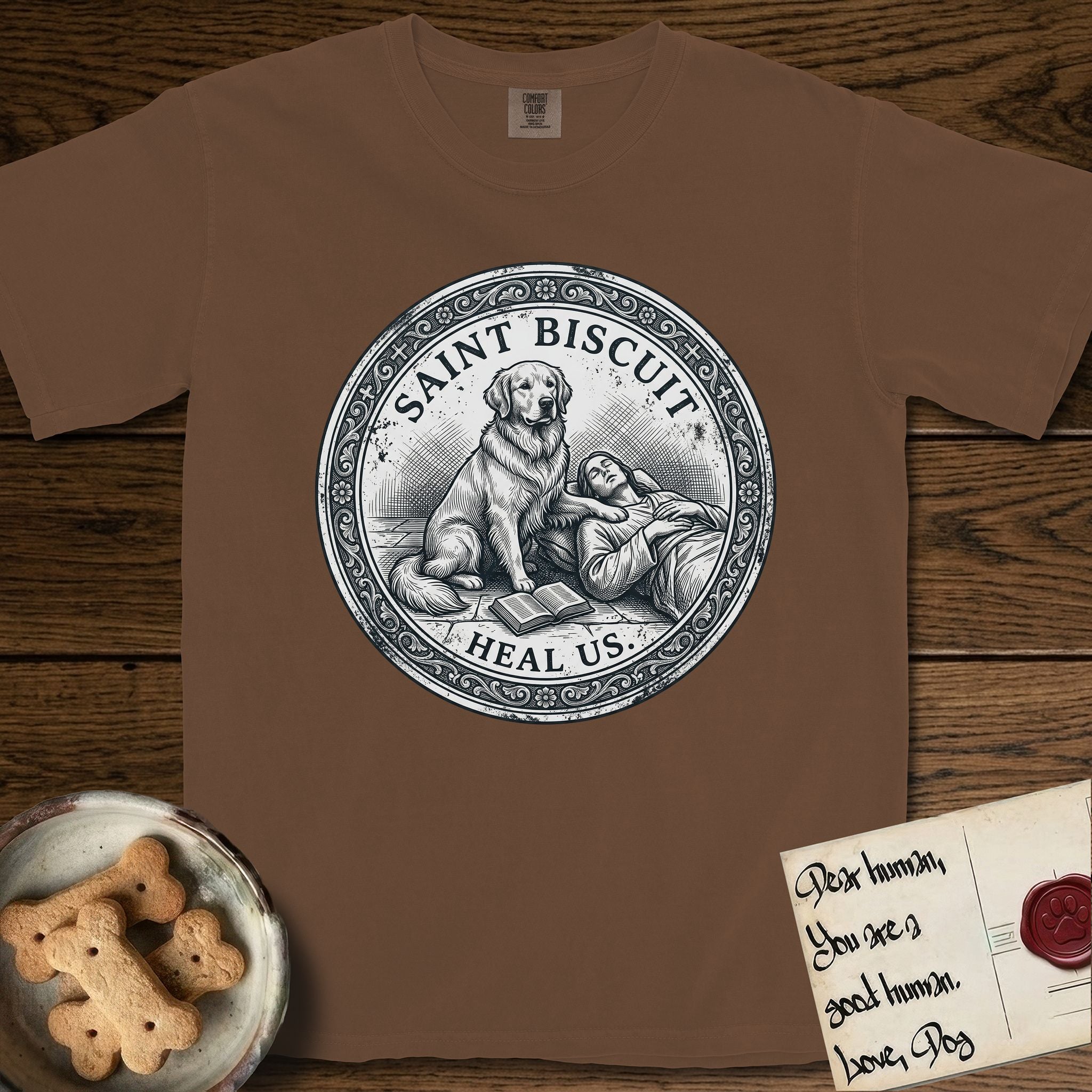 Patron Saint Medallion Saint Biscuit T-Shirt flat lay in Espresso, featuring a vintage engraving coin medallion of a haloed Golden Retriever with a devotee resting against her and an open book, with the petition HEAL US.