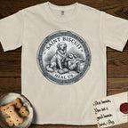 Patron Saint Medallion Saint Biscuit T-Shirt flat lay in Ivory, featuring a vintage engraving coin medallion of a haloed Golden Retriever with a devotee resting against her and an open book, with the petition HEAL US.