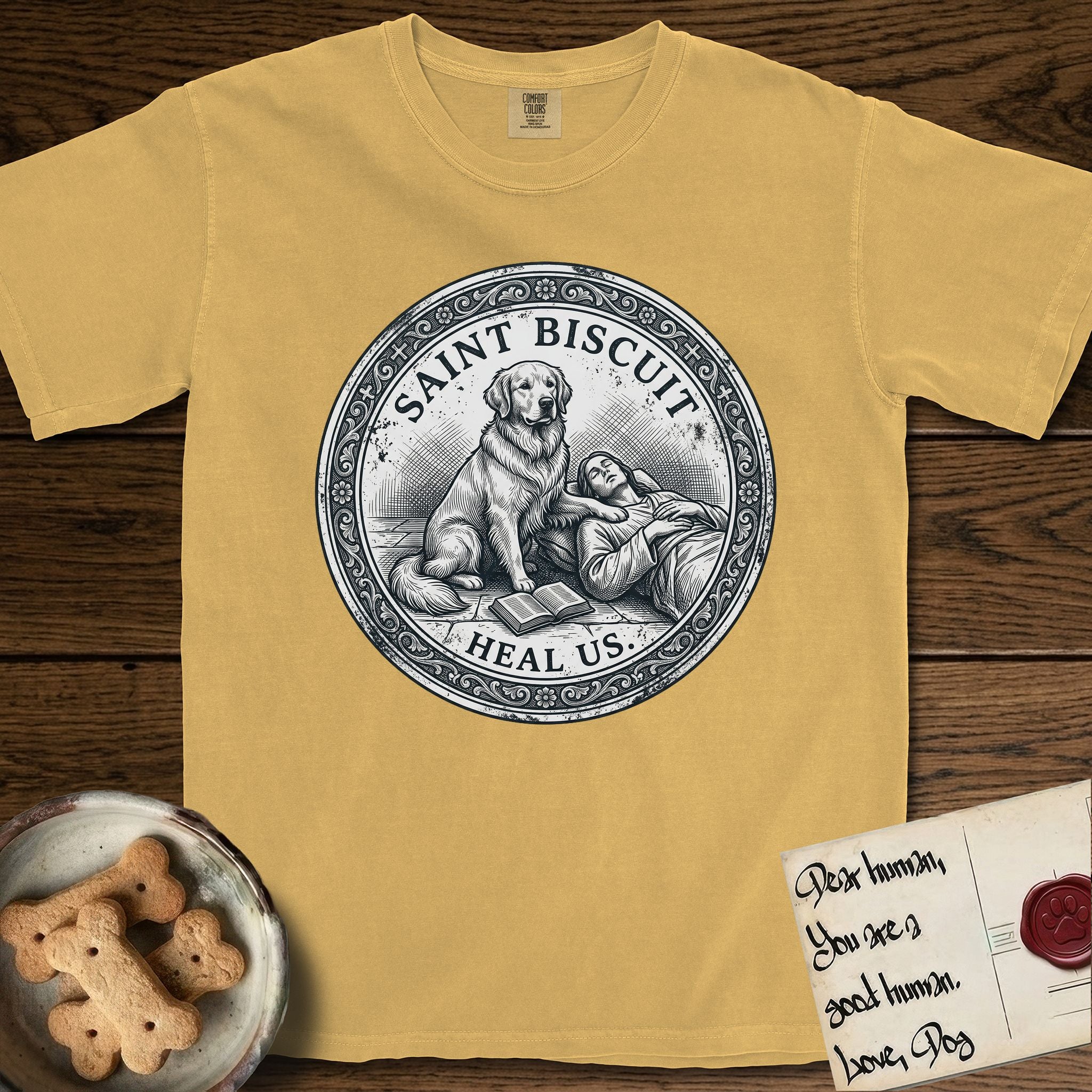 Patron Saint Medallion Saint Biscuit T-Shirt flat lay in Mustard, featuring a vintage engraving coin medallion of a haloed Golden Retriever with a devotee resting against her and an open book, with the petition HEAL US.