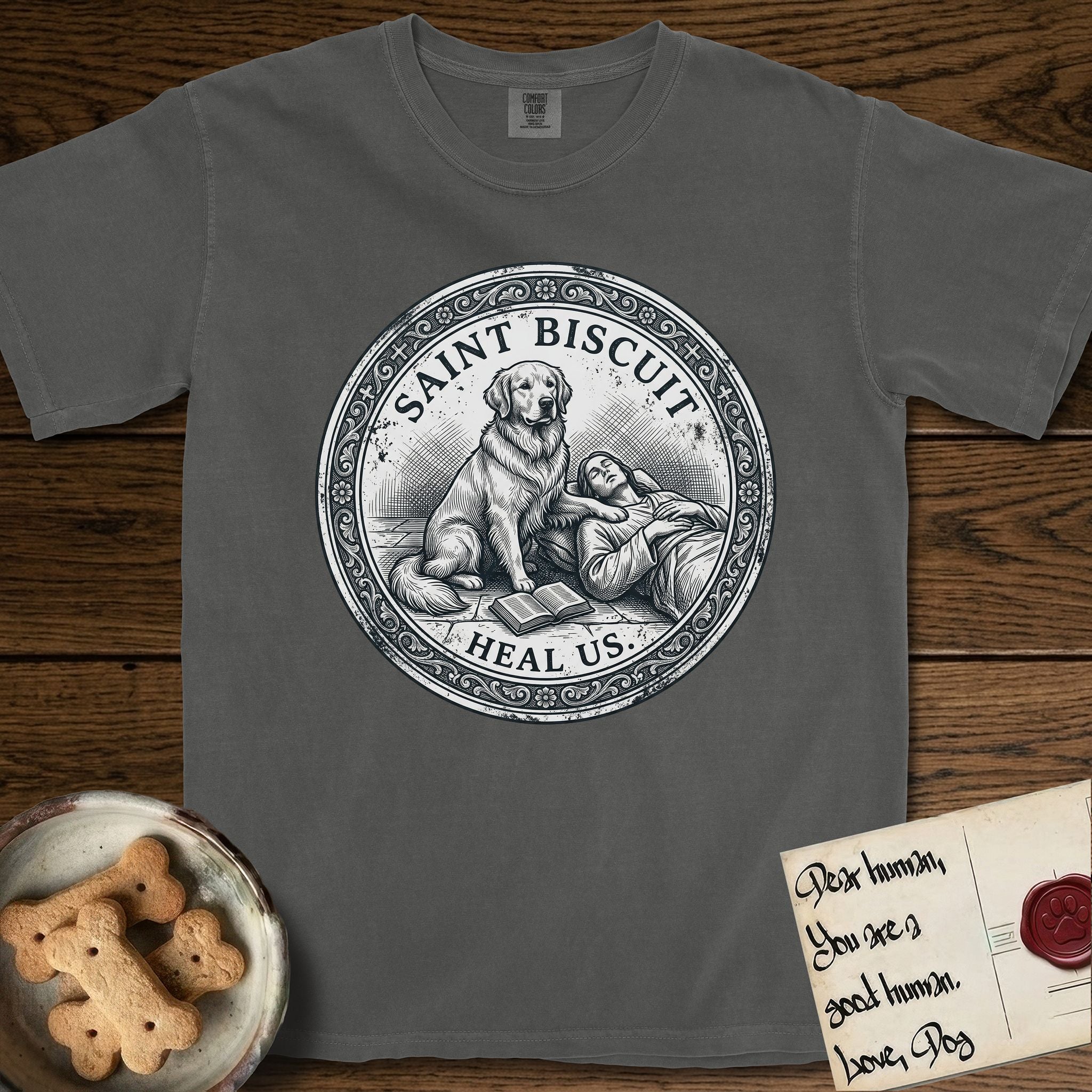 Patron Saint Medallion Saint Biscuit T-Shirt flat lay in Pepper, featuring a vintage engraving coin medallion of a haloed Golden Retriever with a devotee resting against her and an open book, with the petition HEAL US.