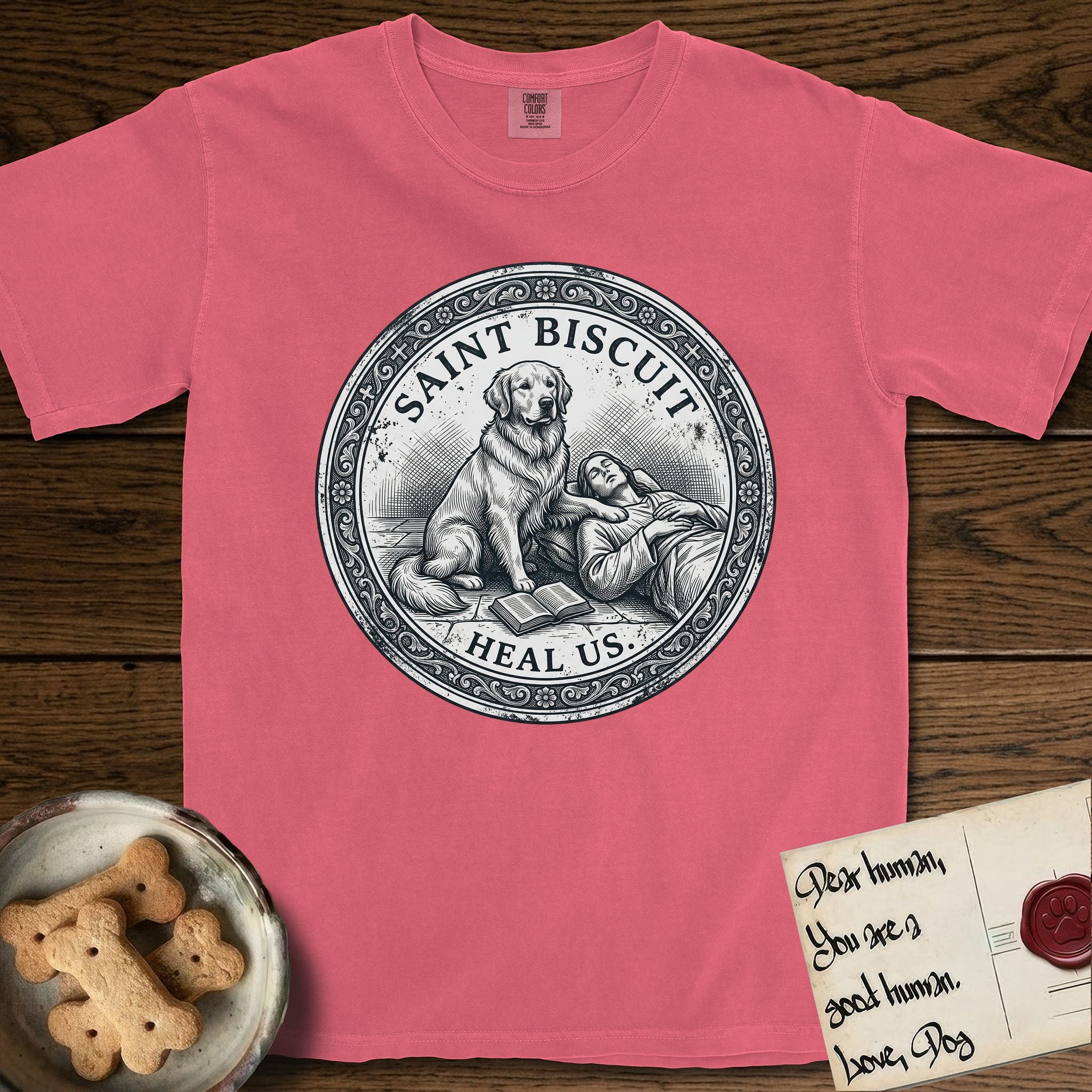 Patron Saint Medallion Saint Biscuit T-Shirt flat lay in Watermelon, featuring a vintage engraving coin medallion of a haloed Golden Retriever with a devotee resting against her and an open book, with the petition HEAL US.