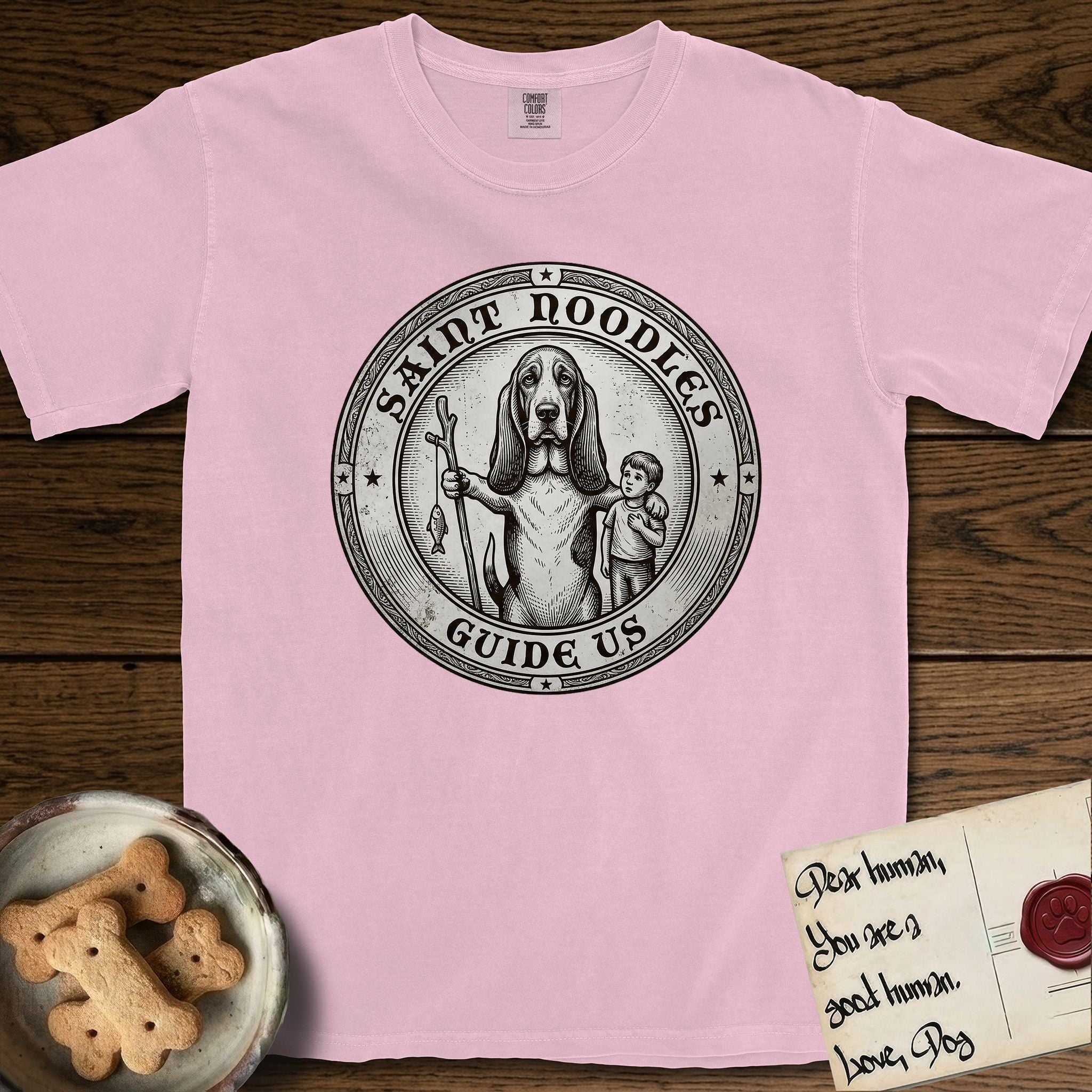 Patron Saint Medallion Saint Noodles T-Shirt flat lay in Blossom, featuring a vintage engraving coin medallion of a haloed Basset Hound with a staff and a child at her side, with the petition GUIDE US.
