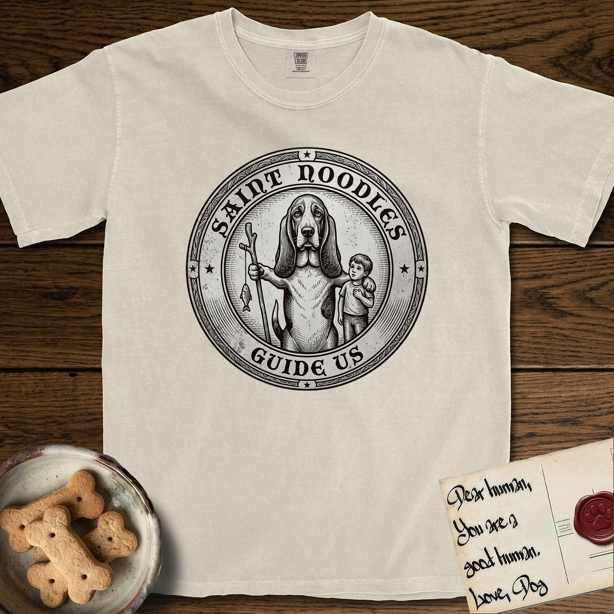 Patron Saint Medallion Saint Noodles T-Shirt flat lay in Ivory, featuring a vintage engraving coin medallion of a haloed Basset Hound with a staff and a child at her side, with the petition GUIDE US.