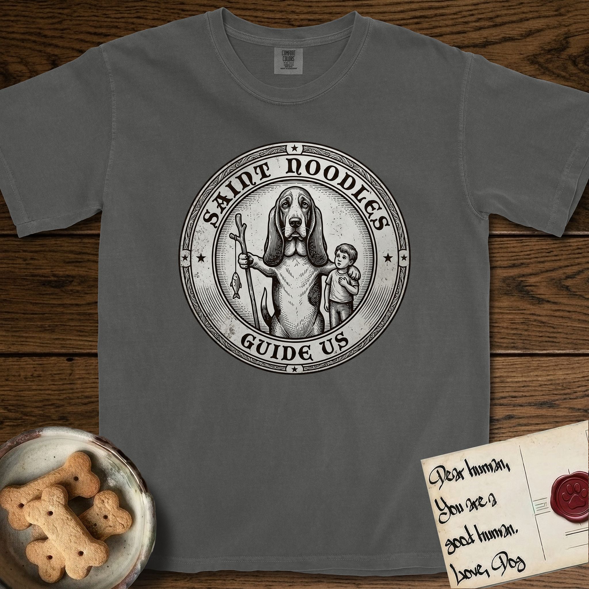Patron Saint Medallion Saint Noodles T-Shirt flat lay in Pepper, featuring a vintage engraving coin medallion of a haloed Basset Hound with a staff and a child at her side, with the petition GUIDE US.