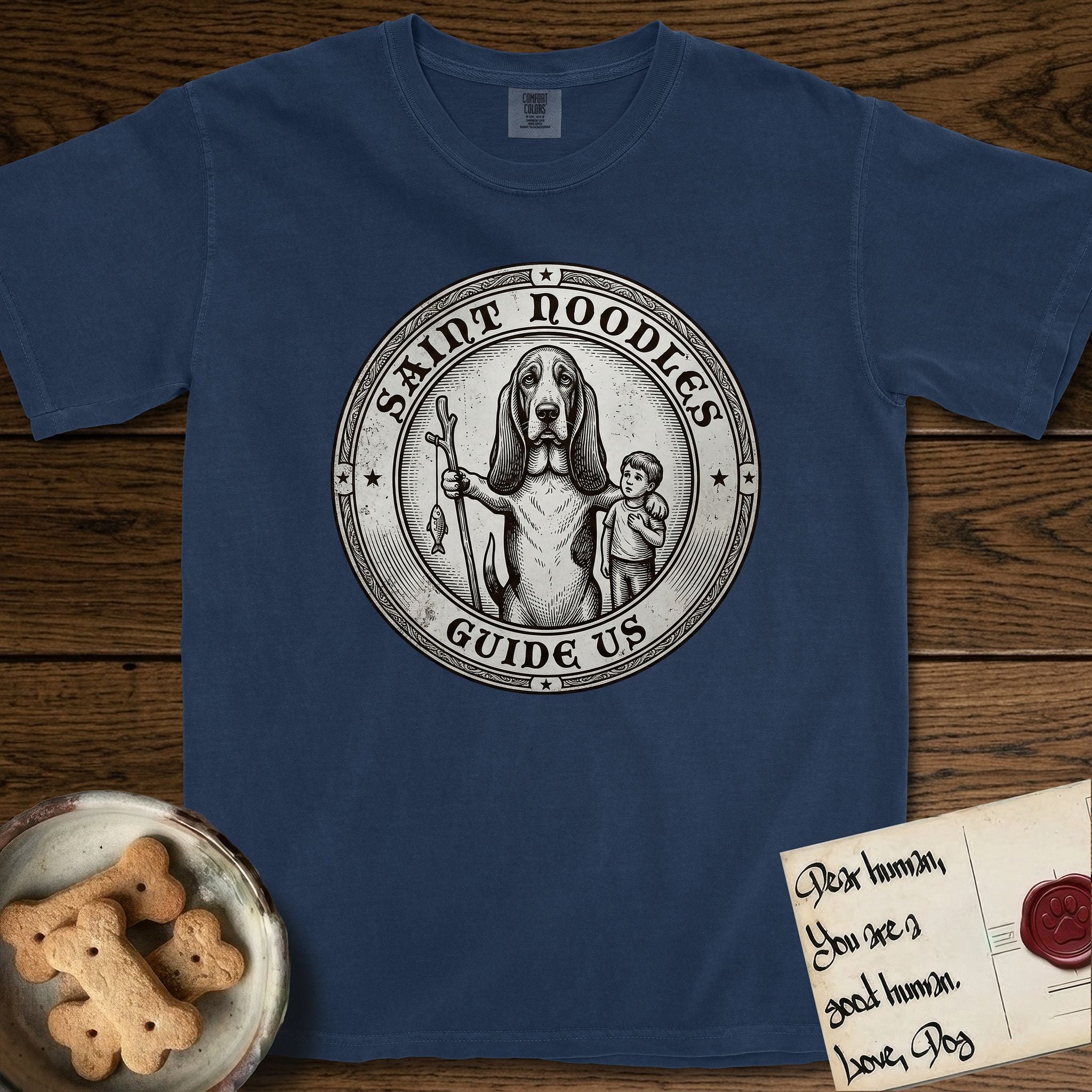 Patron Saint Medallion Saint Noodles T-Shirt flat lay in True Navy, featuring a vintage engraving coin medallion of a haloed Basset Hound with a staff and a child at her side, with the petition GUIDE US.