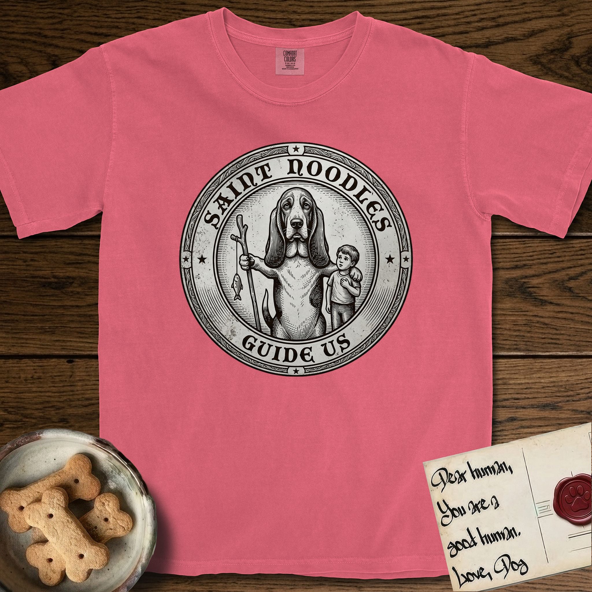 Patron Saint Medallion Saint Noodles T-Shirt flat lay in Watermelon, featuring a vintage engraving coin medallion of a haloed Basset Hound with a staff and a child at her side, with the petition GUIDE US.