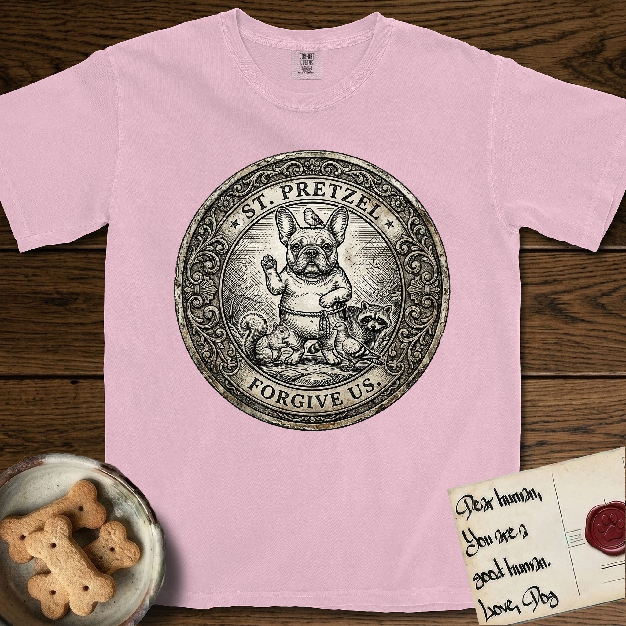 Patron Saint Medallion Saint Pretzel T-Shirt flat lay in Blossom, featuring an ornate vintage engraving coin medallion of a haloed French Bulldog raising one paw, a small bird on his head and woodland animals at his feet, with the petition FORGIVE US.