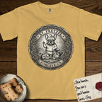 Patron Saint Medallion Saint Pretzel T-Shirt flat lay in Mustard, featuring an ornate vintage engraving coin medallion of a haloed French Bulldog raising one paw, a small bird on his head and woodland animals at his feet, with the petition FORGIVE US.