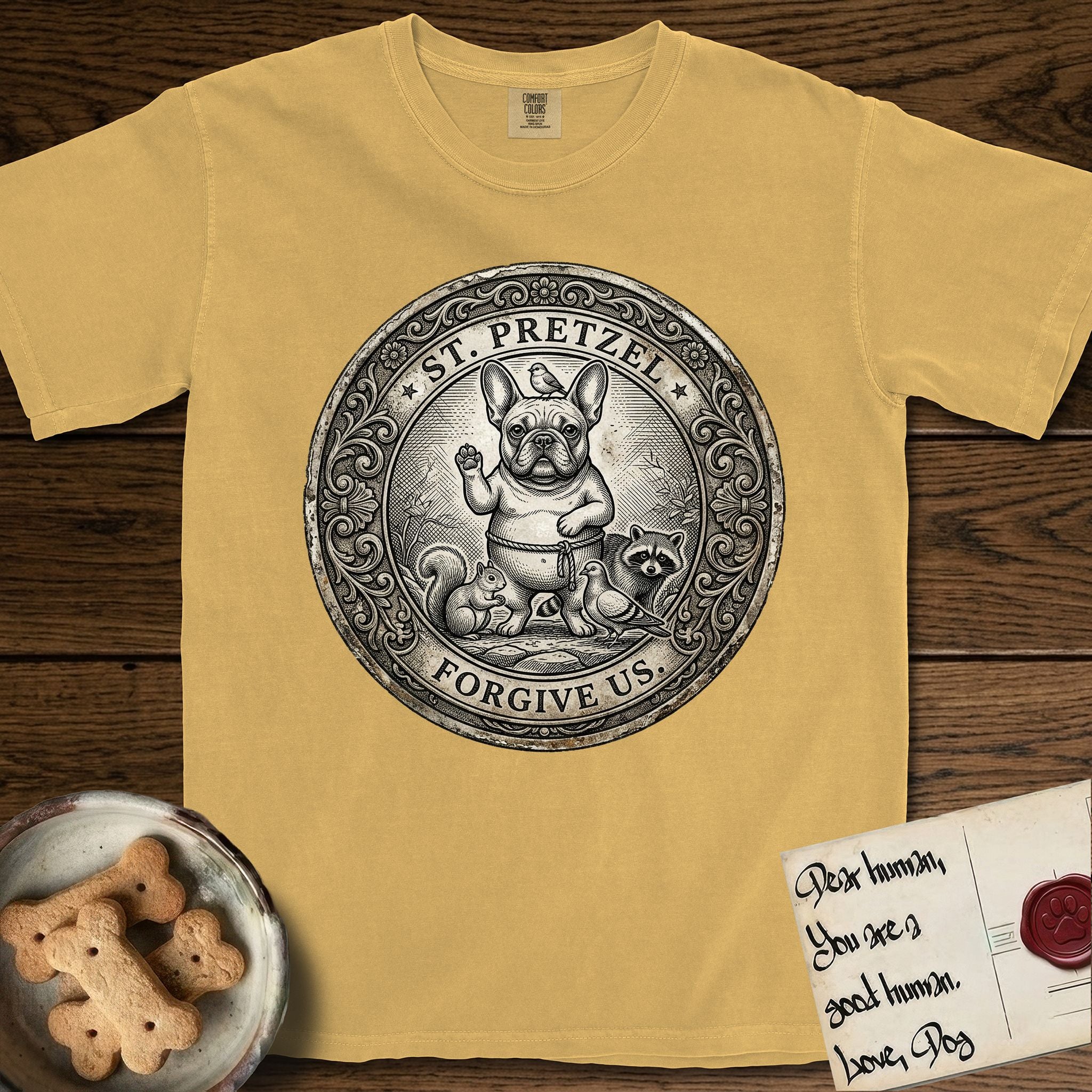 Patron Saint Medallion Saint Pretzel T-Shirt flat lay in Mustard, featuring an ornate vintage engraving coin medallion of a haloed French Bulldog raising one paw, a small bird on his head and woodland animals at his feet, with the petition FORGIVE US.