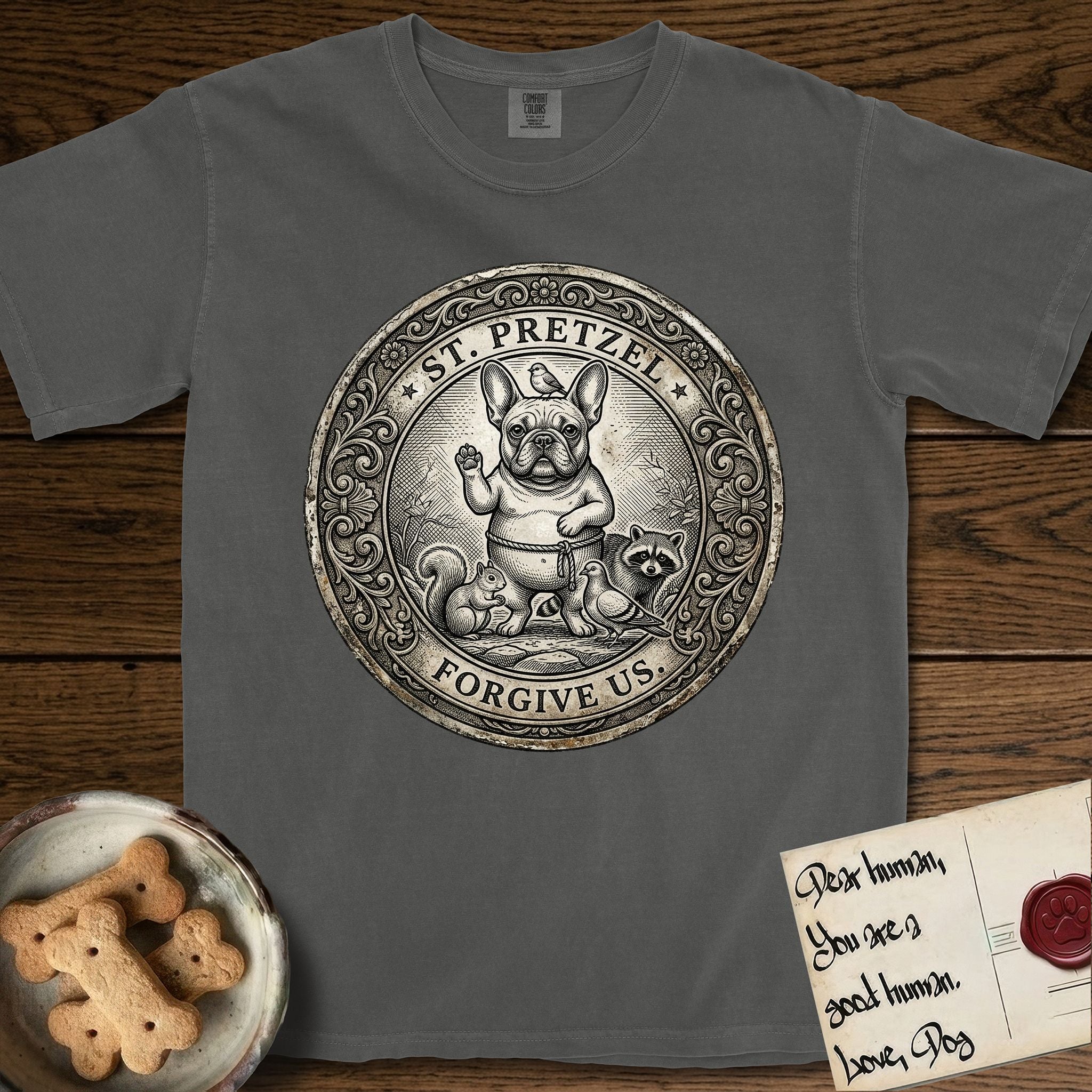 Patron Saint Medallion Saint Pretzel T-Shirt flat lay in Pepper, featuring an ornate vintage engraving coin medallion of a haloed French Bulldog raising one paw, a small bird on his head and woodland animals at his feet, with the petition FORGIVE US.