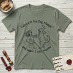Rejoice in the Dog Goldendoodle T-Shirt flat lay in Sage, featuring a sparse pen-and-ink illustration of a girl and her Goldendoodle dancing together, fur flying, both heads thrown back.