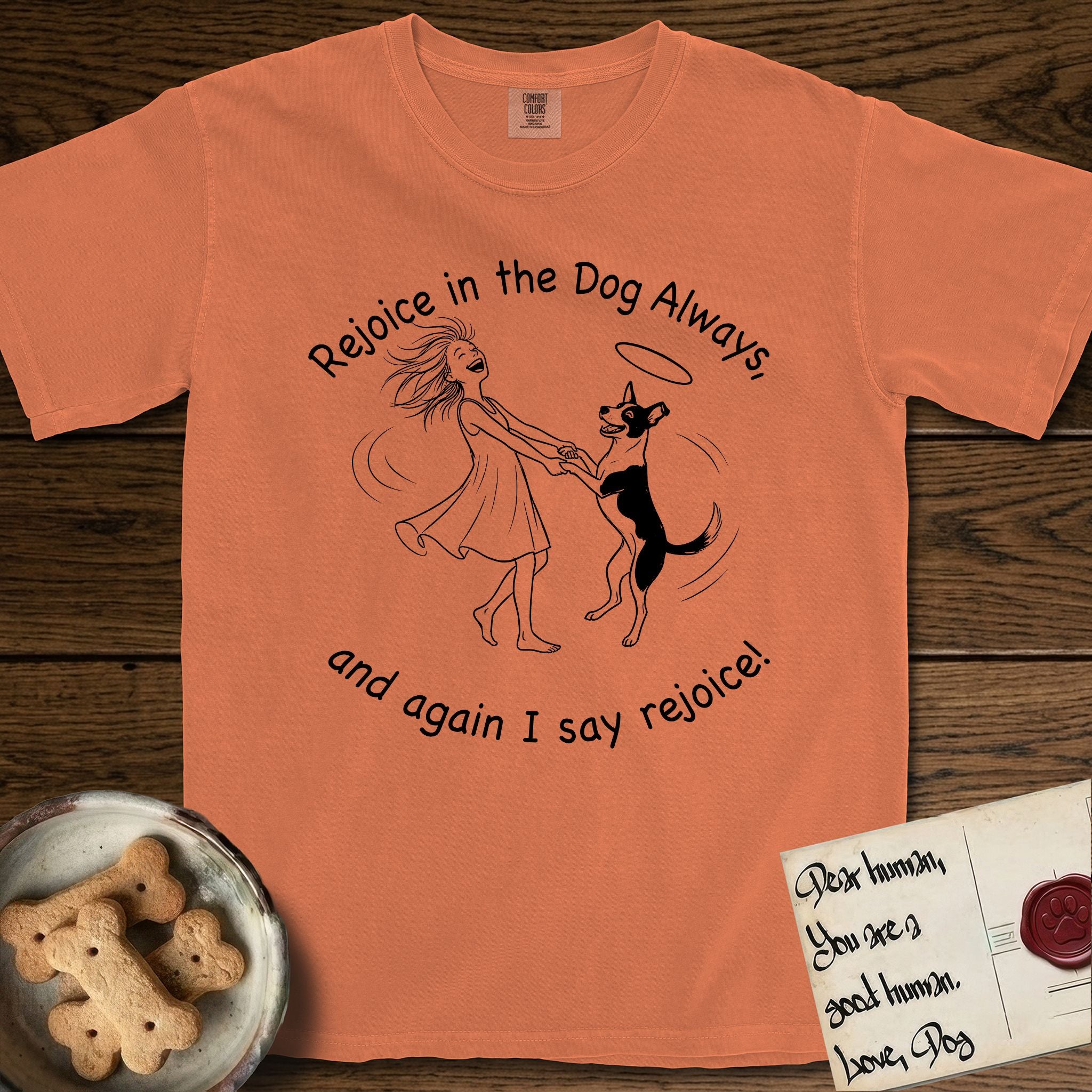 Rejoice in the Dog Lobo T-Shirt flat lay in Melon, featuring a sparse pen-and-ink illustration of a girl and her haloed Dog Lobo spinning together, hands and paws clasped.