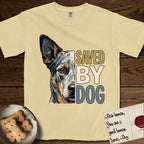Saved by Dog Australian Cattle Dog T-Shirt flat lay in Butter, featuring half an Australian Cattle Dog's face in distressed vintage screen print, blue-speckled with tan points and one amber eye, cropped down the centerline beside bold stacked block lettering.