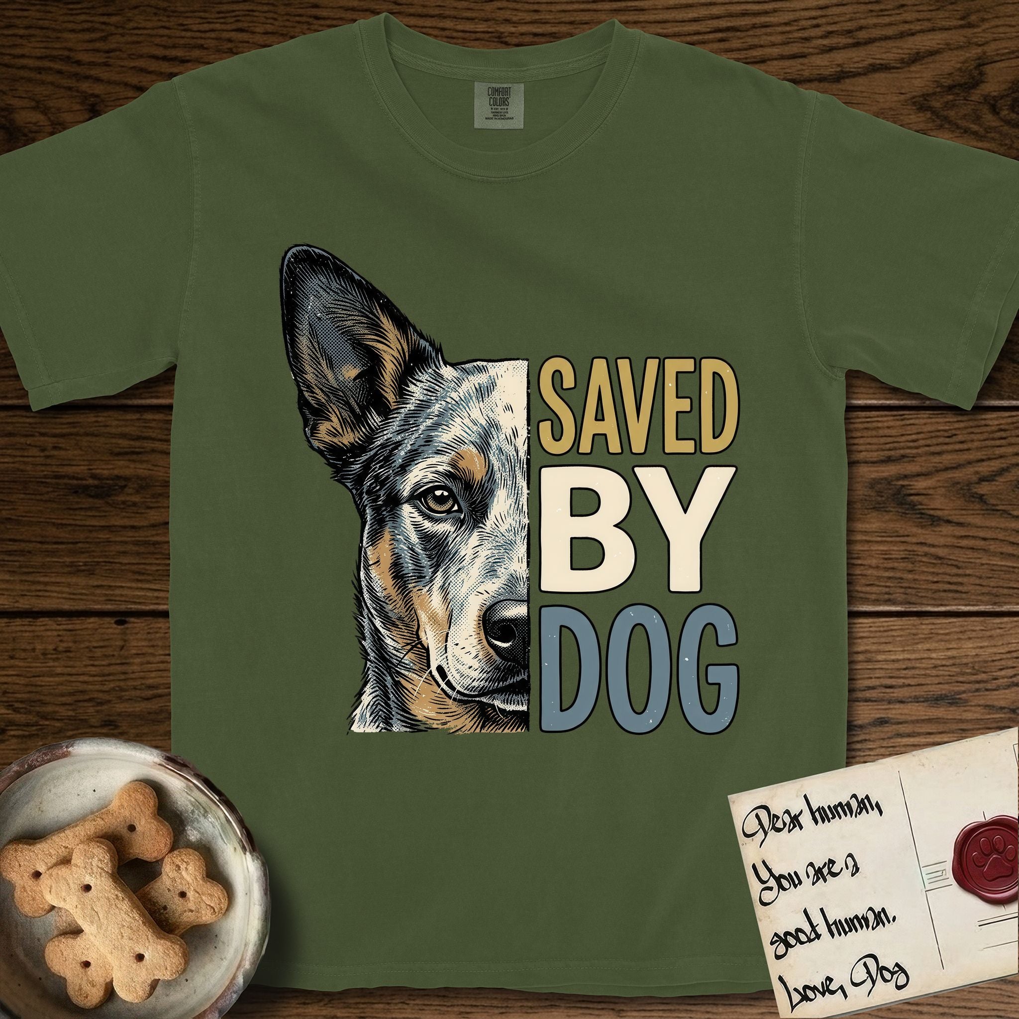 Saved by Dog Australian Cattle Dog T-Shirt flat lay in Hemp, featuring half an Australian Cattle Dog's face in distressed vintage screen print, blue-speckled with tan points and one amber eye, cropped down the centerline beside bold stacked block lettering.