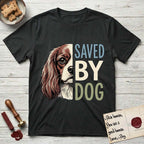 Saved by Dog Cavalier T-Shirt flat lay in Black, featuring half a chestnut-and-white Cavalier's face in distressed vintage screen print, one long feathered ear and a large soft eye, cropped down the centerline beside bold stacked block lettering.