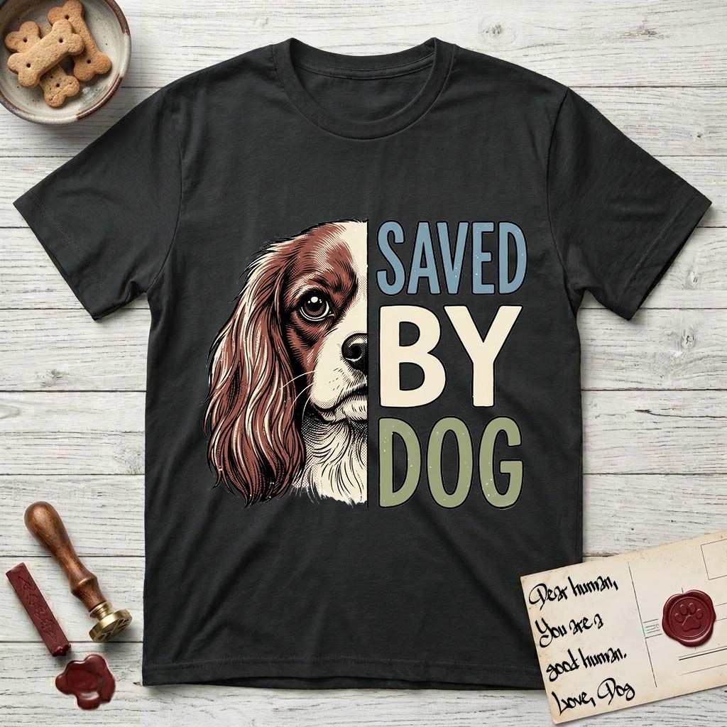 Saved by Dog Cavalier T-Shirt flat lay in Black, featuring half a chestnut-and-white Cavalier's face in distressed vintage screen print, one long feathered ear and a large soft eye, cropped down the centerline beside bold stacked block lettering.