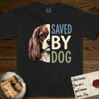 Saved by Dog Cavalier T-Shirt flat lay in Black, featuring half a chestnut-and-white Cavalier's face in distressed vintage screen print, one long feathered ear and a large soft eye, cropped down the centerline beside bold stacked block lettering.