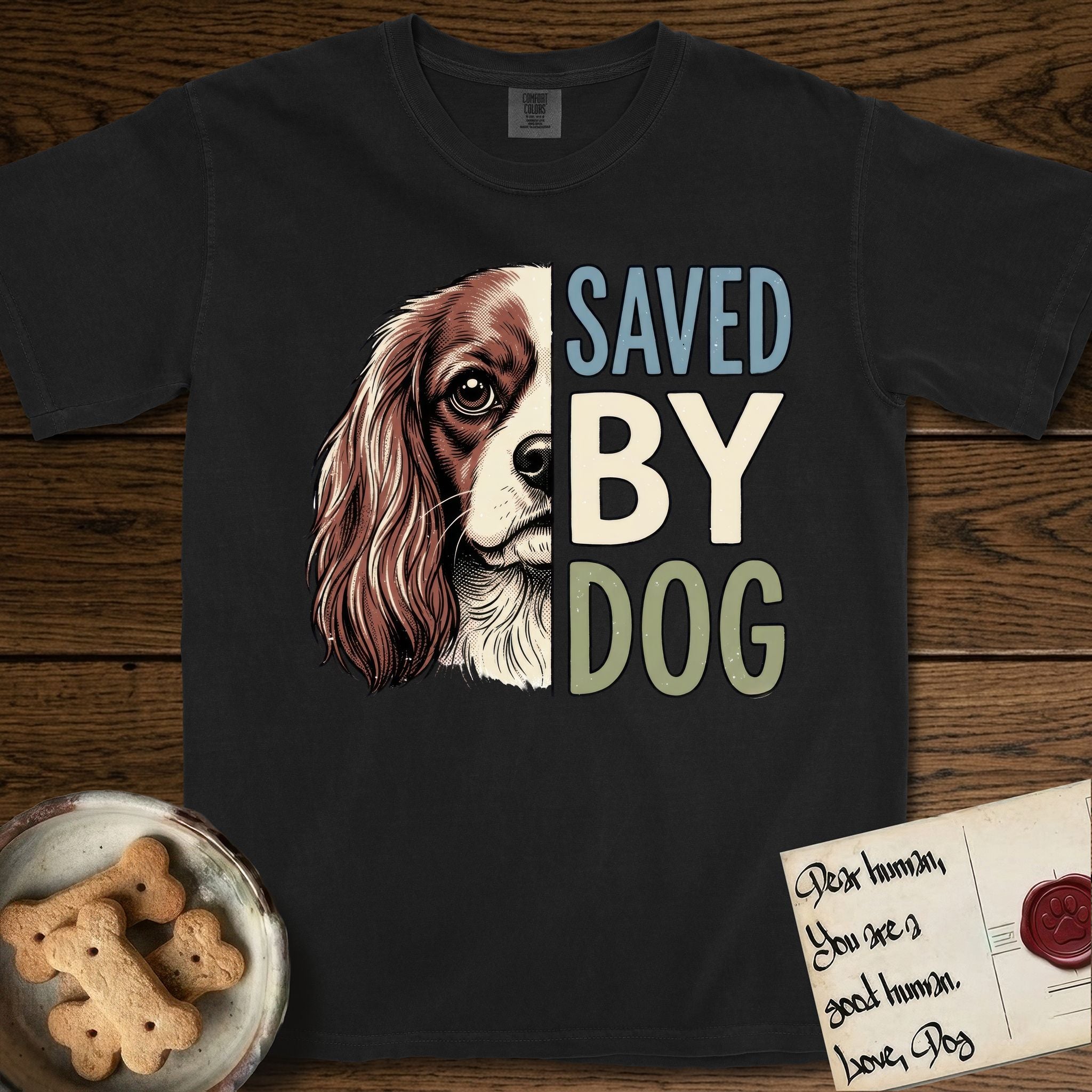 Saved by Dog Cavalier T-Shirt flat lay in Black, featuring half a chestnut-and-white Cavalier's face in distressed vintage screen print, one long feathered ear and a large soft eye, cropped down the centerline beside bold stacked block lettering.