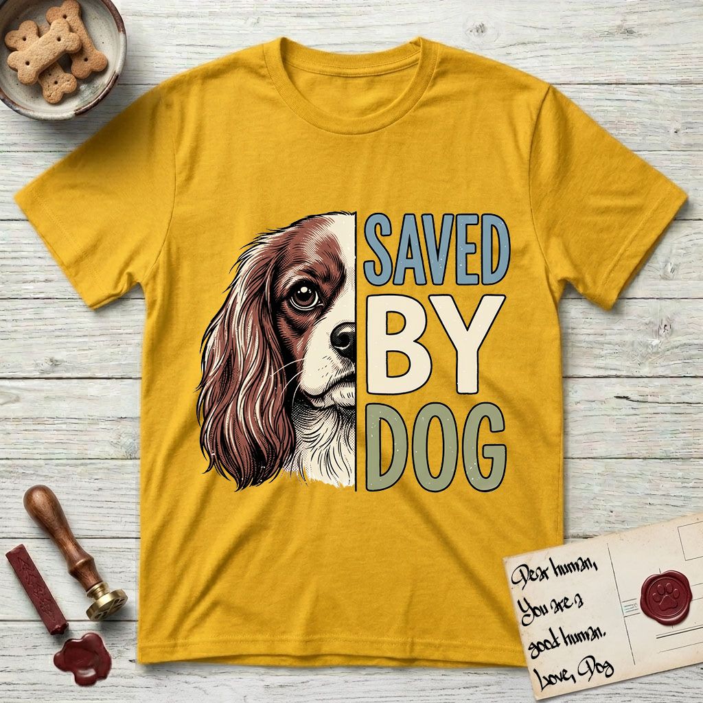 Saved by Dog Cavalier T-Shirt flat lay in Daisy, featuring half a chestnut-and-white Cavalier's face in distressed vintage screen print, one long feathered ear and a large soft eye, cropped down the centerline beside bold stacked block lettering.