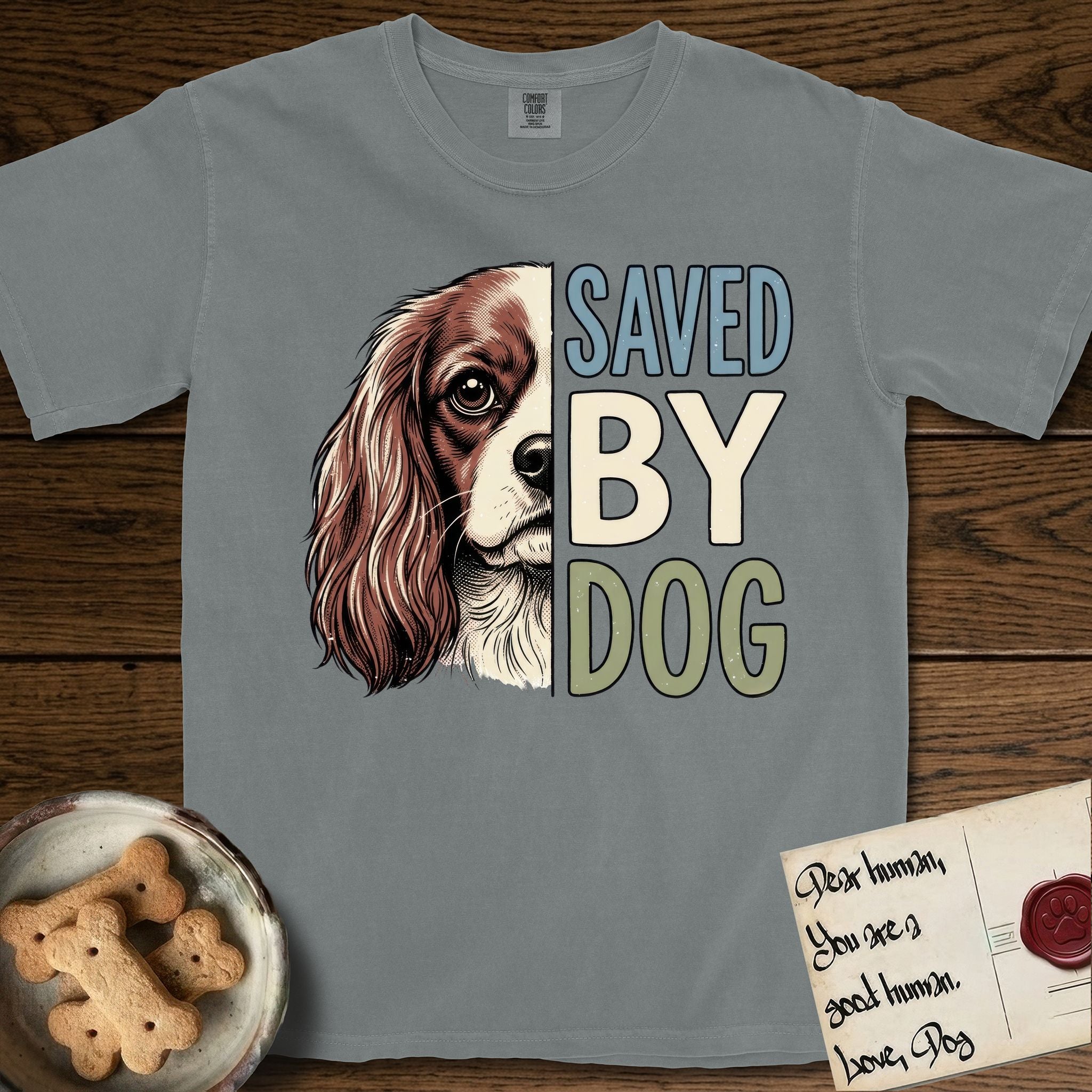 Saved by Dog Cavalier T-Shirt flat lay in Granite, featuring half a chestnut-and-white Cavalier's face in distressed vintage screen print, one long feathered ear and a large soft eye, cropped down the centerline beside bold stacked block lettering.