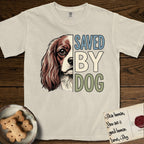 Saved by Dog Cavalier T-Shirt flat lay in Ivory, featuring half a chestnut-and-white Cavalier's face in distressed vintage screen print, one long feathered ear and a large soft eye, cropped down the centerline beside bold stacked block lettering.