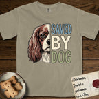 Saved by Dog Cavalier T-Shirt flat lay in Khaki, featuring half a chestnut-and-white Cavalier's face in distressed vintage screen print, one long feathered ear and a large soft eye, cropped down the centerline beside bold stacked block lettering.
