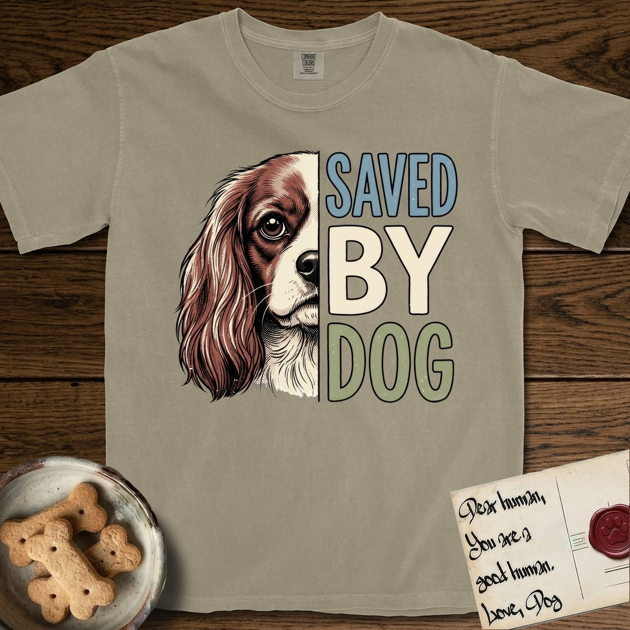 Saved by Dog Cavalier T-Shirt flat lay in Khaki, featuring half a chestnut-and-white Cavalier's face in distressed vintage screen print, one long feathered ear and a large soft eye, cropped down the centerline beside bold stacked block lettering.
