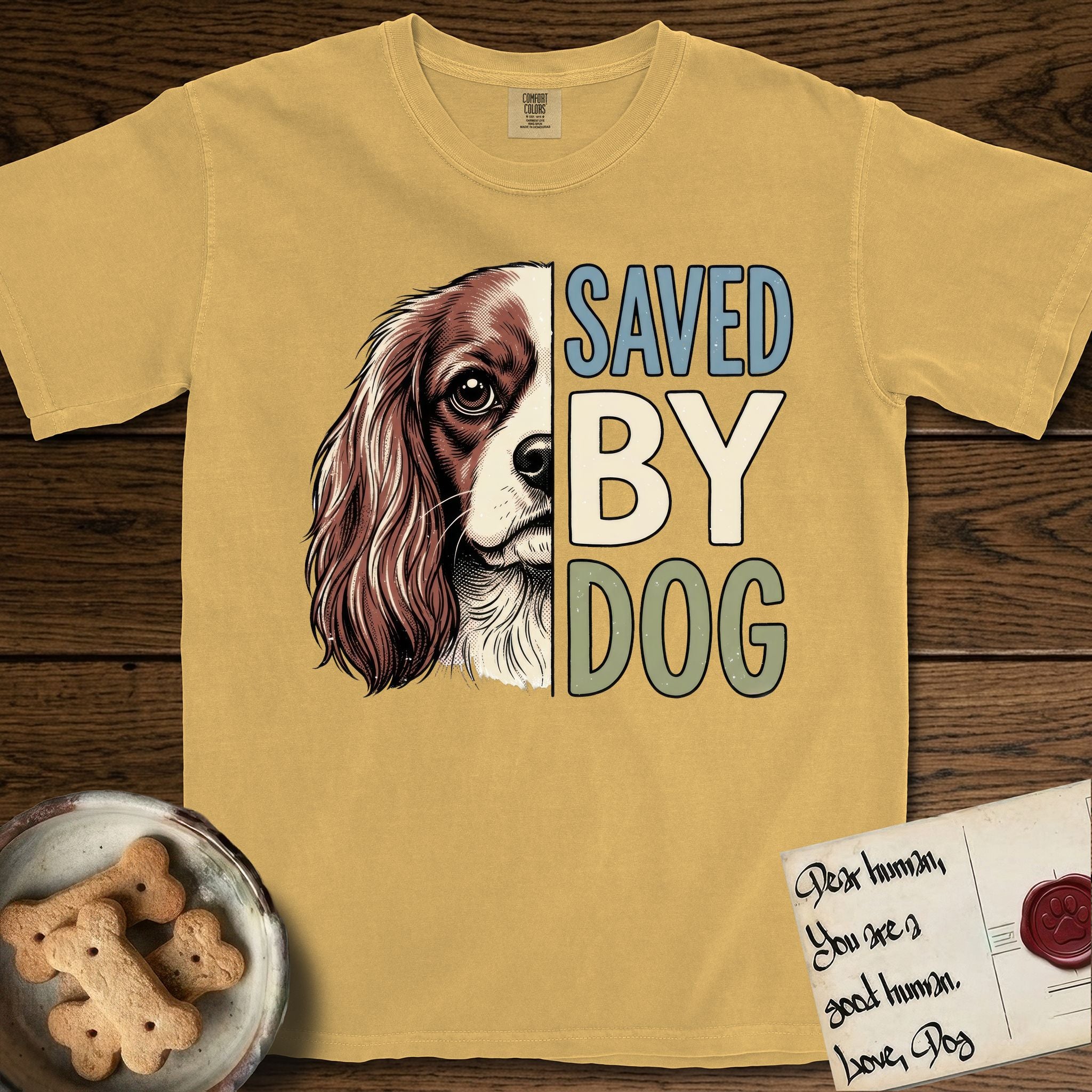 Saved by Dog Cavalier T-Shirt flat lay in Mustard, featuring half a chestnut-and-white Cavalier's face in distressed vintage screen print, one long feathered ear and a large soft eye, cropped down the centerline beside bold stacked block lettering.