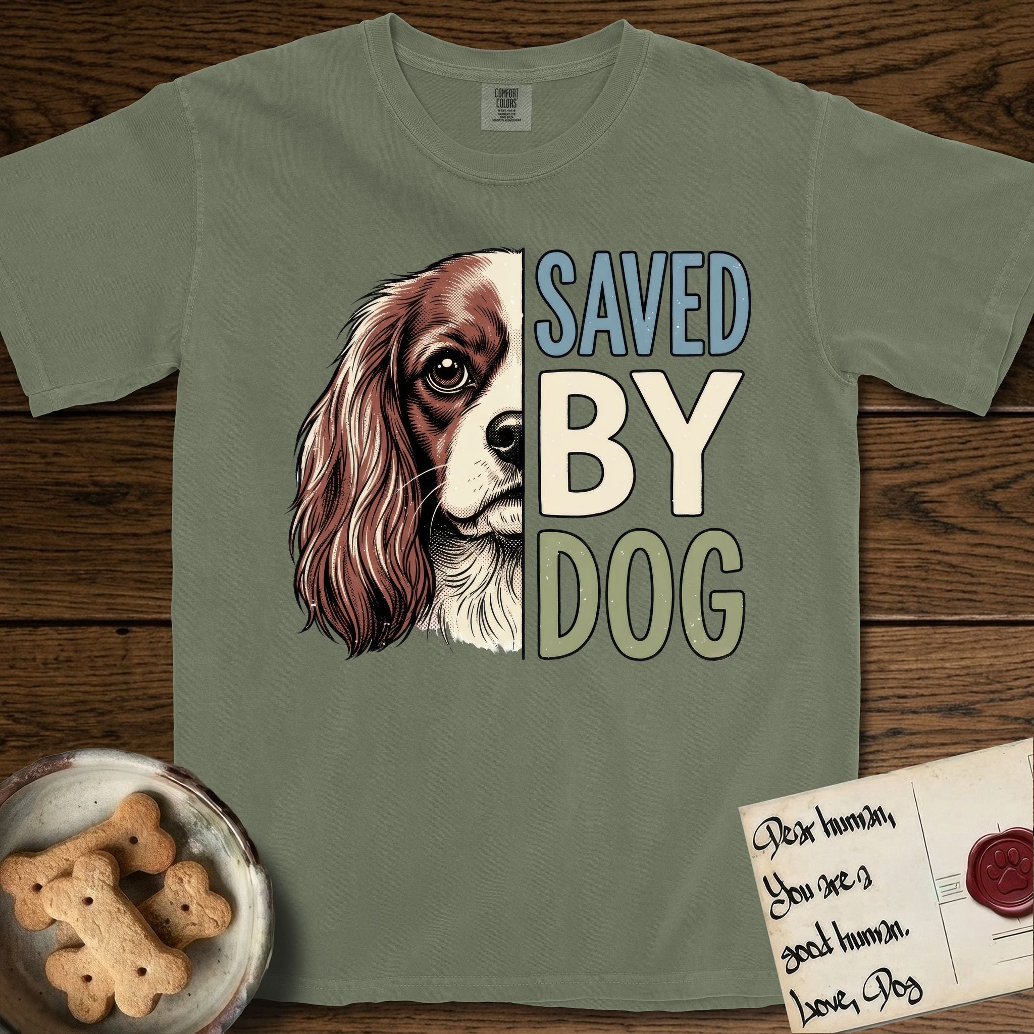 Saved by Dog Cavalier T-Shirt flat lay in Sage, featuring half a chestnut-and-white Cavalier's face in distressed vintage screen print, one long feathered ear and a large soft eye, cropped down the centerline beside bold stacked block lettering.