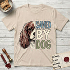 Saved by Dog Cavalier T-Shirt flat lay in Sand, featuring half a chestnut-and-white Cavalier's face in distressed vintage screen print, one long feathered ear and a large soft eye, cropped down the centerline beside bold stacked block lettering.