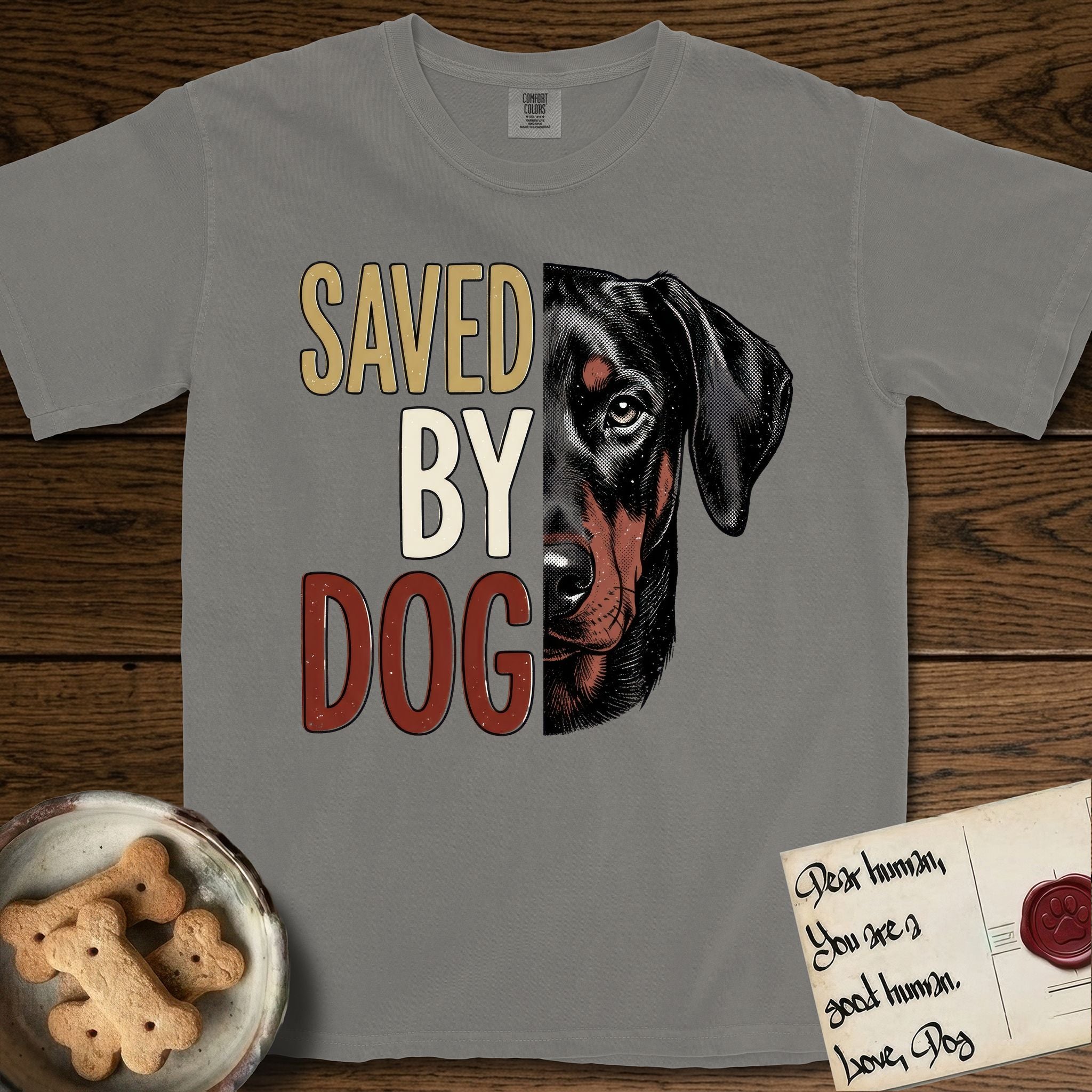 Saved by Dog Doberman T-Shirt flat lay in Grey, featuring half a black-and-rust Doberman's face in distressed vintage screen print, natural uncropped ears and a steady eye, cropped down the centerline beside bold stacked block lettering.