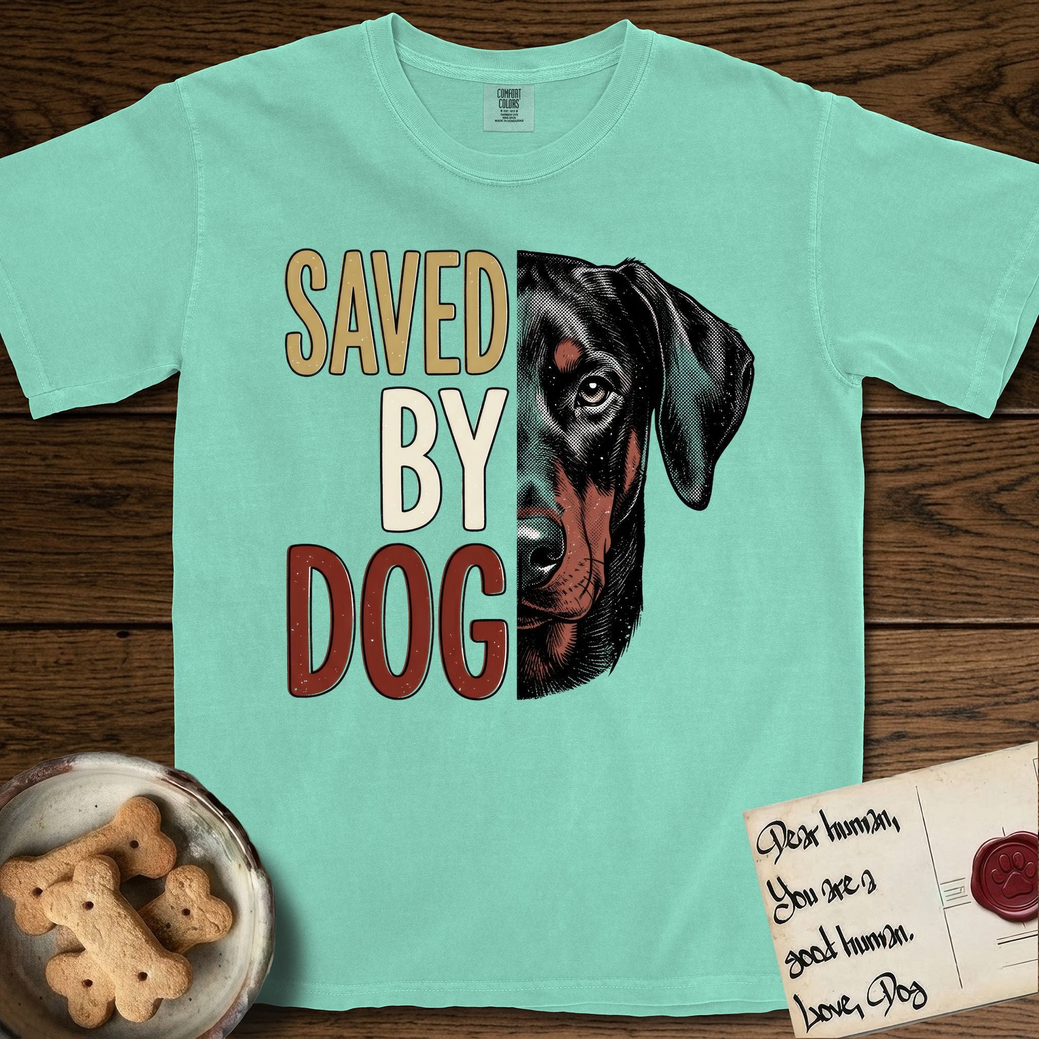 Saved by Dog Doberman T-Shirt flat lay in Island Reef, featuring half a black-and-rust Doberman's face in distressed vintage screen print, natural uncropped ears and a steady eye, cropped down the centerline beside bold stacked block lettering.