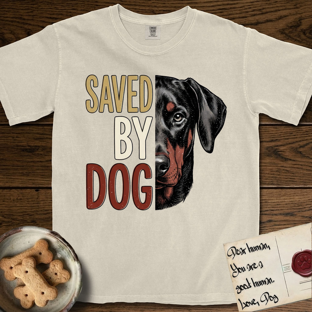 Saved by Dog Doberman T-Shirt flat lay in Ivory, featuring half a black-and-rust Doberman's face in distressed vintage screen print, natural uncropped ears and a steady eye, cropped down the centerline beside bold stacked block lettering.