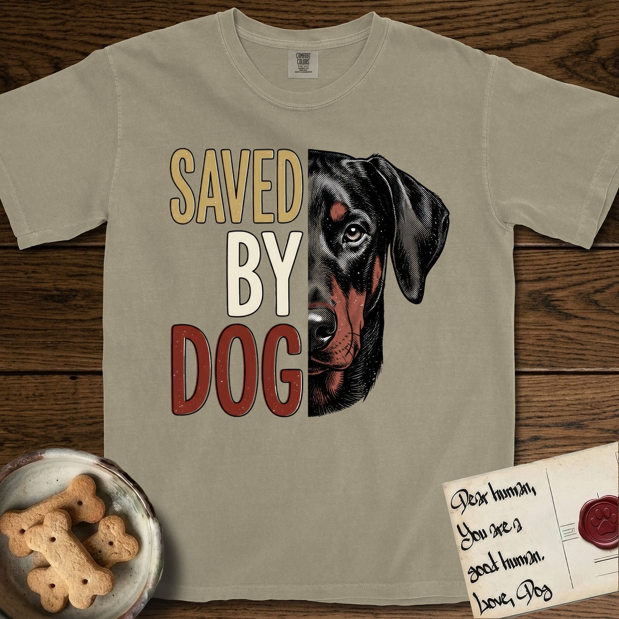 Saved by Dog Doberman T-Shirt flat lay in Khaki, featuring half a black-and-rust Doberman's face in distressed vintage screen print, natural uncropped ears and a steady eye, cropped down the centerline beside bold stacked block lettering.