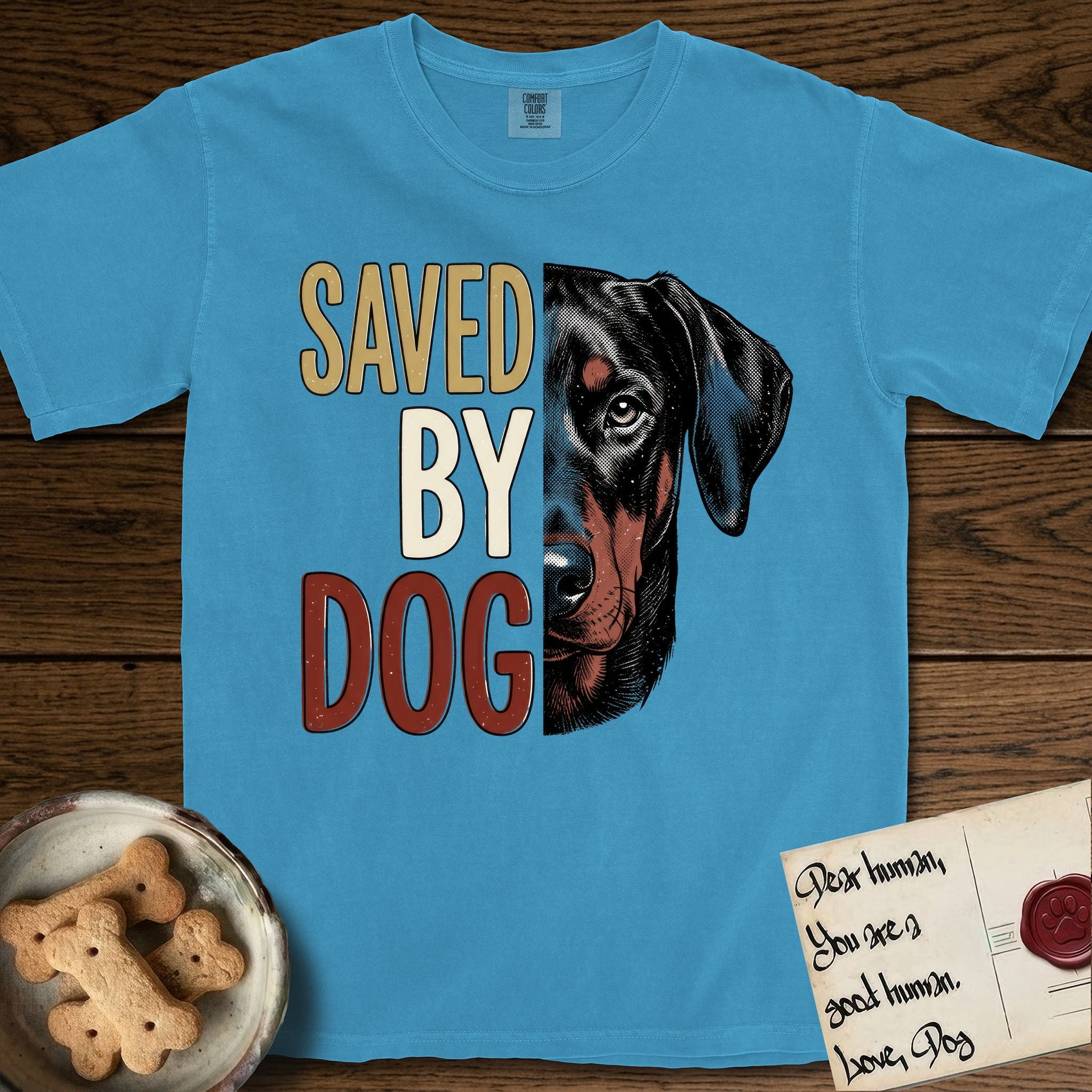 Saved by Dog Doberman T-Shirt flat lay in Royal Caribe, featuring half a black-and-rust Doberman's face in distressed vintage screen print, natural uncropped ears and a steady eye, cropped down the centerline beside bold stacked block lettering.