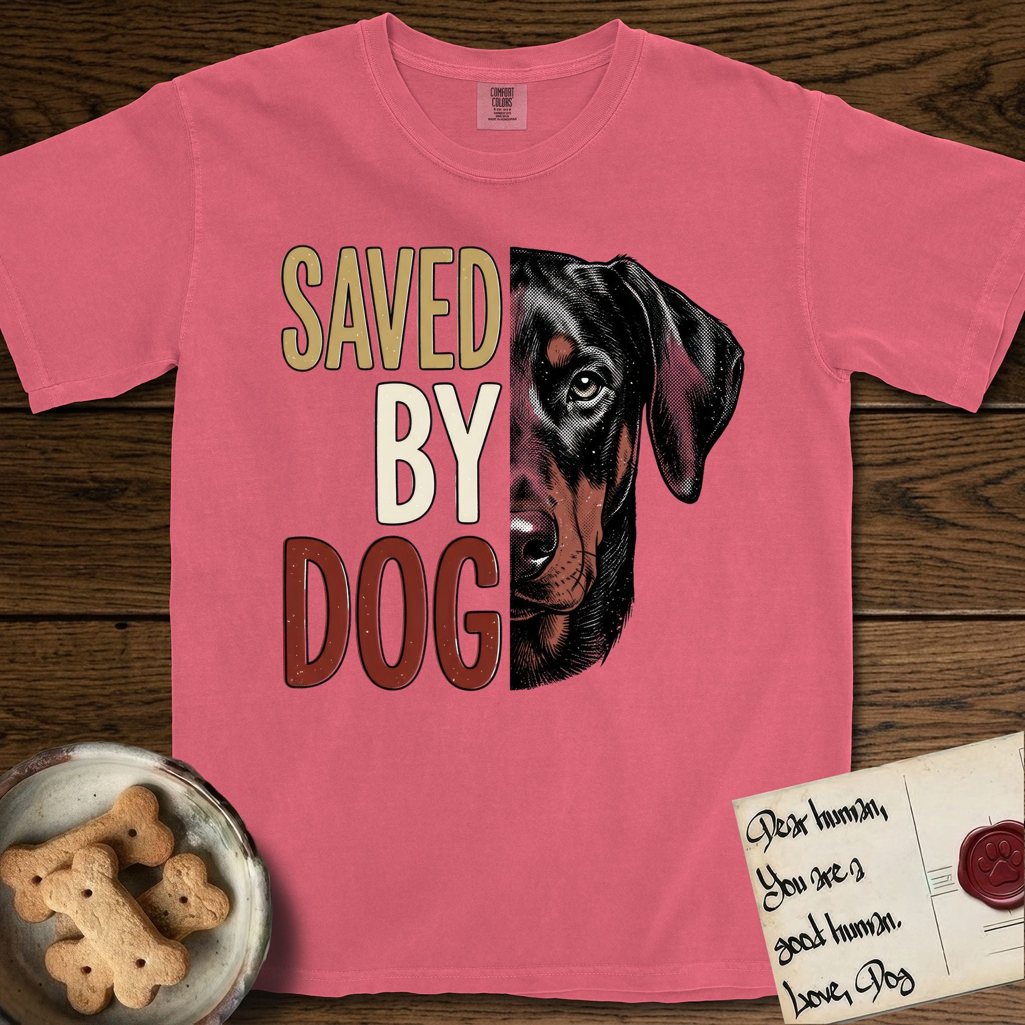 Saved by Dog Doberman T-Shirt flat lay in Watermelon, featuring half a black-and-rust Doberman's face in distressed vintage screen print, natural uncropped ears and a steady eye, cropped down the centerline beside bold stacked block lettering.