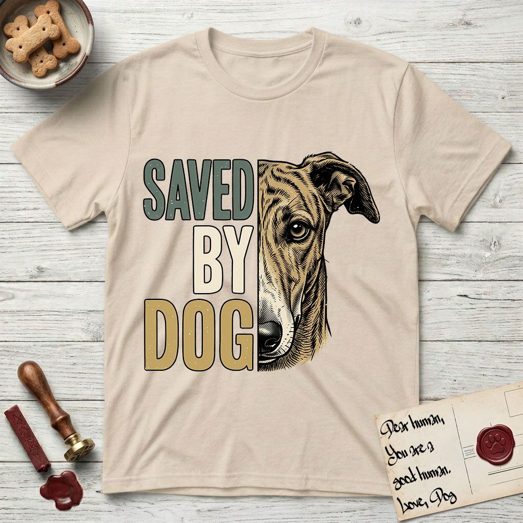 Saved by Dog Galgo T-Shirt flat lay in Sand, featuring half a brindle Galgo's face in distressed vintage screen print, a folded rose ear and a gentle eye, cropped down the centerline beside bold stacked block lettering.