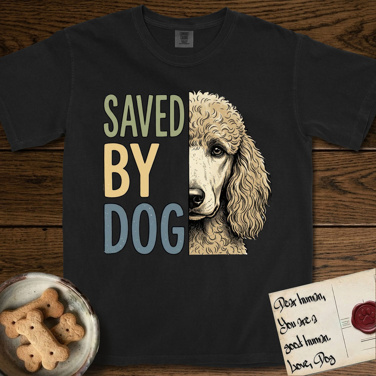 Saved by Dog Poodle T-Shirt flat lay in Black, featuring half an apricot Poodle's face in distressed vintage screen print, soft natural curls and a knowing eye, cropped down the centerline beside bold stacked block lettering.