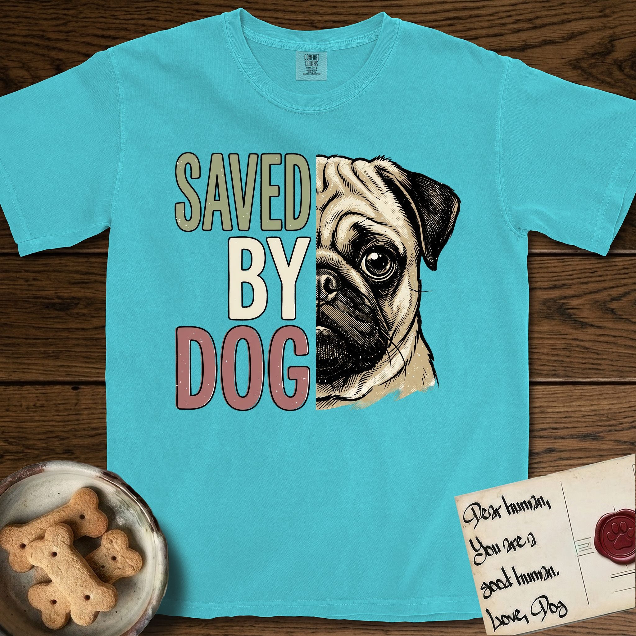 Saved by Dog Pug T-Shirt flat lay in Lagoon Blue, featuring half a fawn Pug's face in distressed vintage screen print, a dark mask and one large round eye, cropped down the centerline beside bold stacked block lettering.