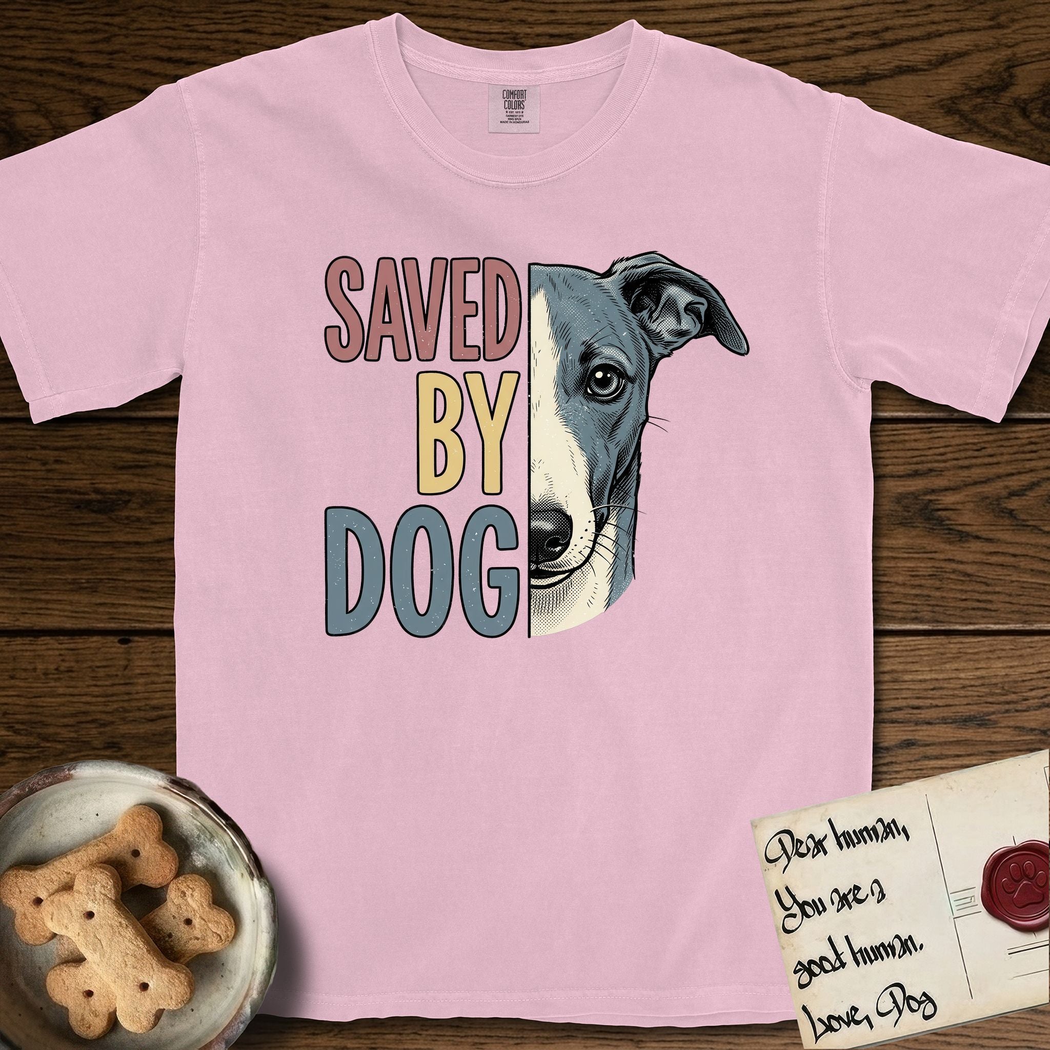 Saved by Dog Whippet T-Shirt flat lay in Blossom, featuring half a blue-grey Whippet's face in distressed vintage screen print, a folded rose ear and a large liquid eye, cropped down the centerline beside bold stacked block lettering.