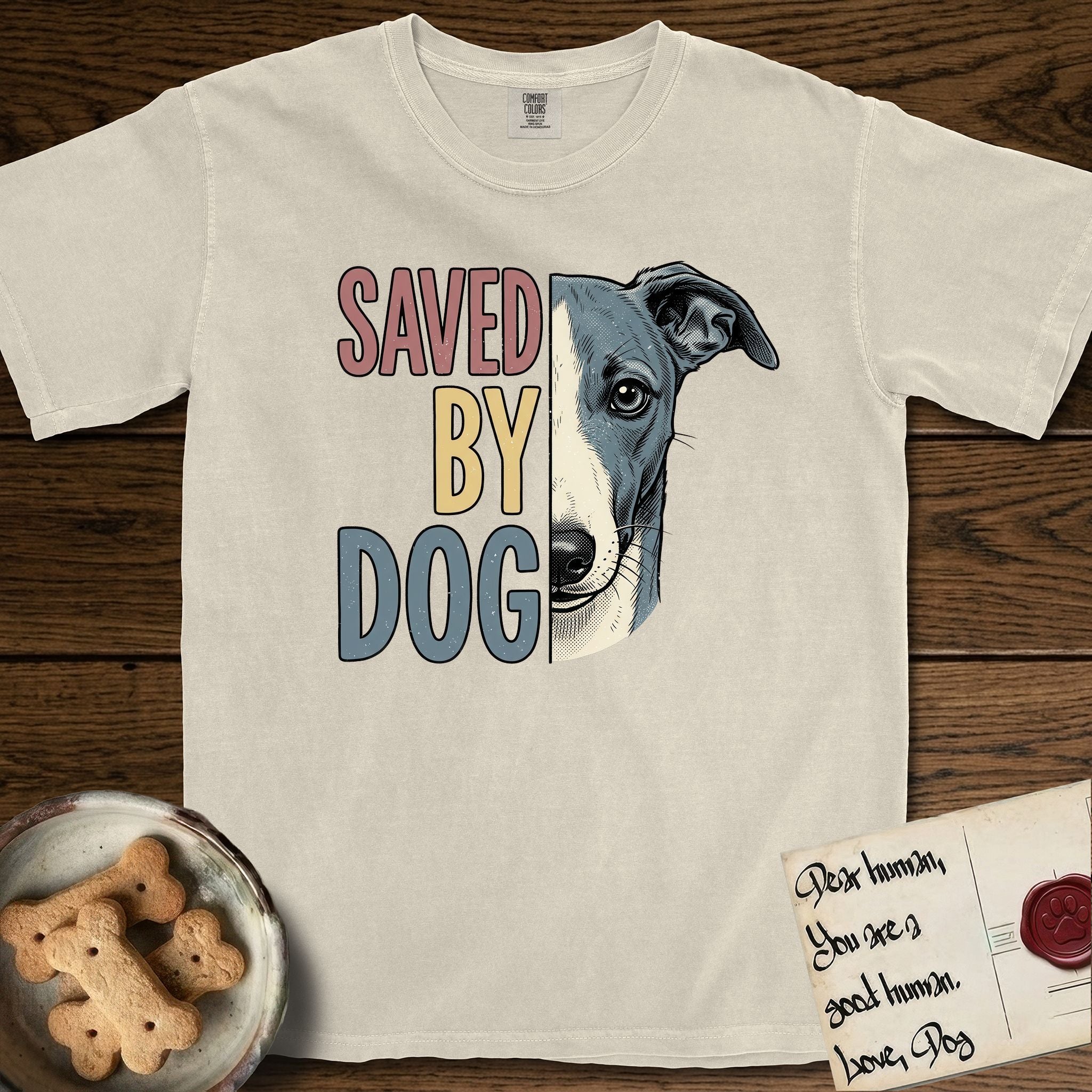 Saved by Dog Whippet T-Shirt flat lay in Ivory, featuring half a blue-grey Whippet's face in distressed vintage screen print, a folded rose ear and a large liquid eye, cropped down the centerline beside bold stacked block lettering.