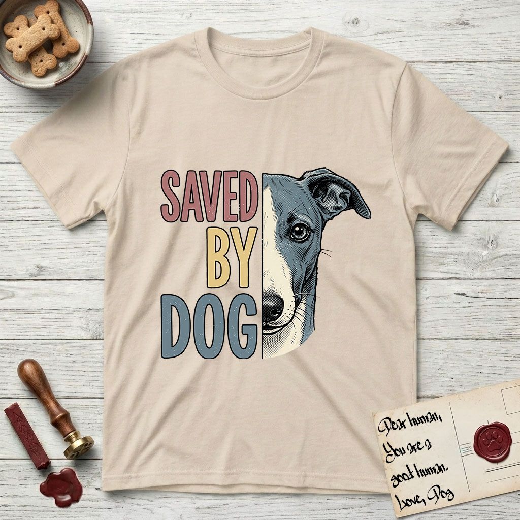 Saved by Dog Whippet T-Shirt flat lay in Sand, featuring half a blue-grey Whippet's face in distressed vintage screen print, a folded rose ear and a large liquid eye, cropped down the centerline beside bold stacked block lettering.