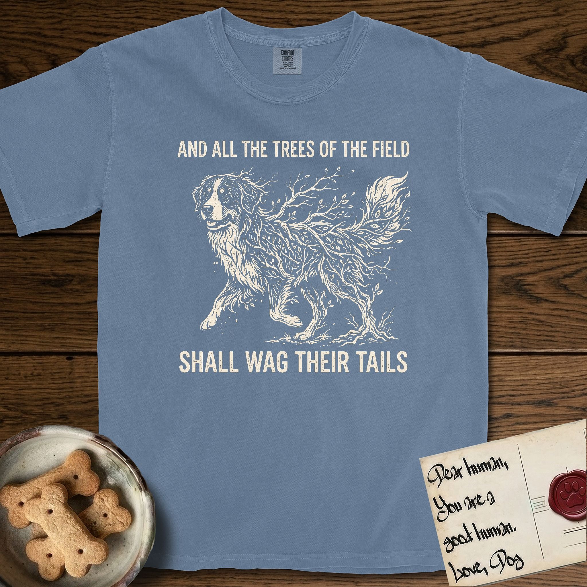 Trees of the Field Bernese Mountain Dog T-Shirt flat lay in Blue Jean, featuring a vintage woodcut illustration of a Bernese Mountain Dog formed of bare windswept winter branches, mid-stride, with the words And All the Trees of the Field Shall Wag Their Tails.