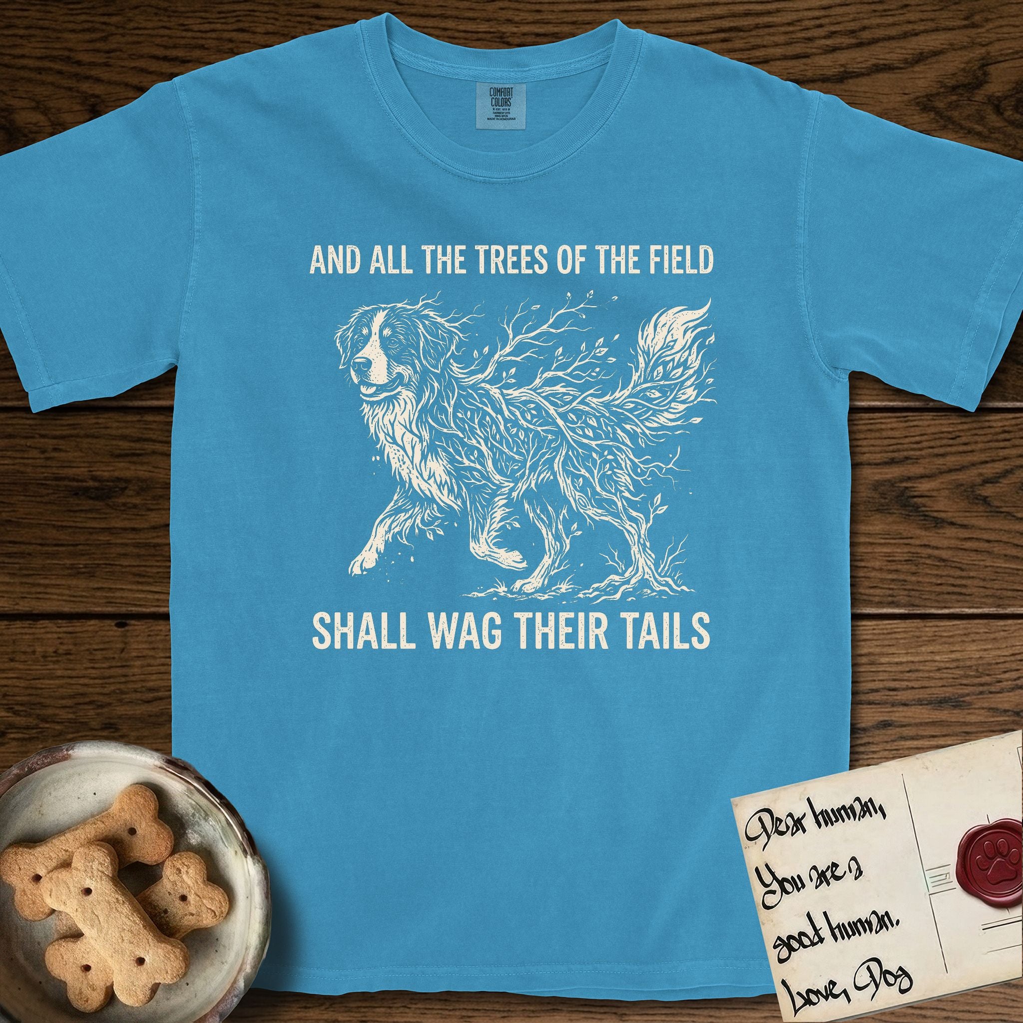 Trees of the Field Bernese Mountain Dog T-Shirt flat lay in Royal Caribe, featuring a vintage woodcut illustration of a Bernese Mountain Dog formed of bare windswept winter branches, mid-stride, with the words And All the Trees of the Field Shall Wag Their Tails.
