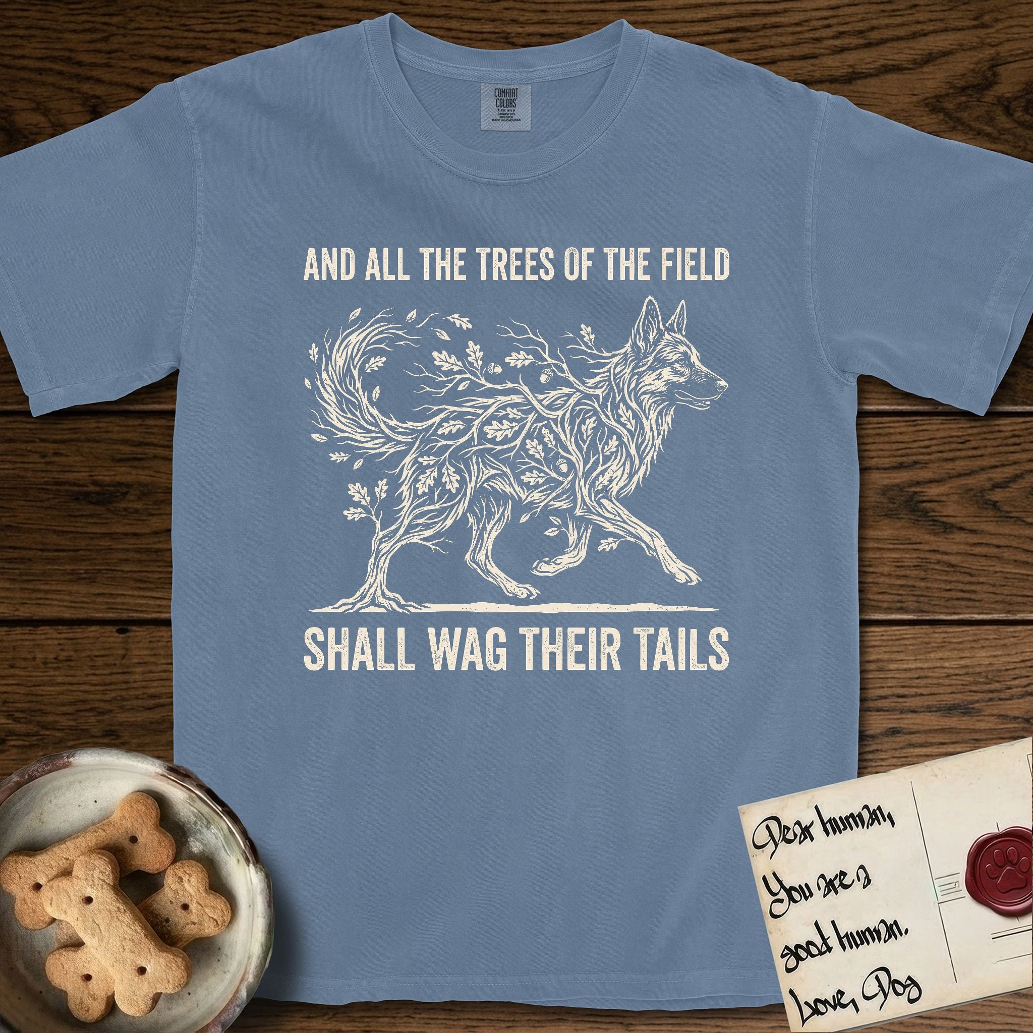 Trees of the Field German Shepherd T-Shirt flat lay in Blue Jean, featuring a vintage woodcut illustration of a German Shepherd formed of oak branches with acorns, mid-stride, with the words And All the Trees of the Field Shall Wag Their Tails.