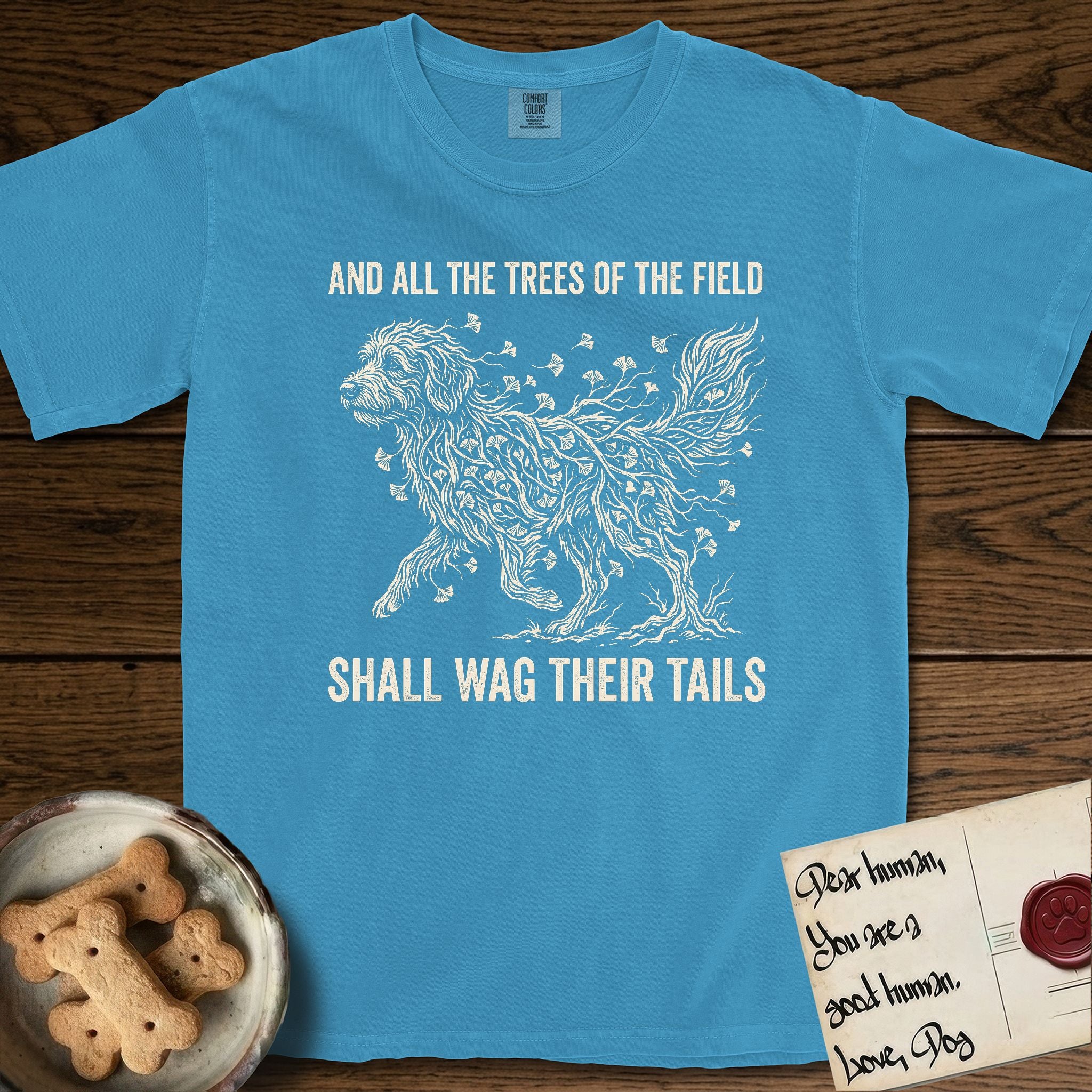 Trees of the Field Goldendoodle T-Shirt flat lay in Royal Caribe, featuring a vintage woodcut illustration of a Goldendoodle formed of ginkgo branches with fan-shaped leaves, mid-stride, with the words And All the Trees of the Field Shall Wag Their Tails.