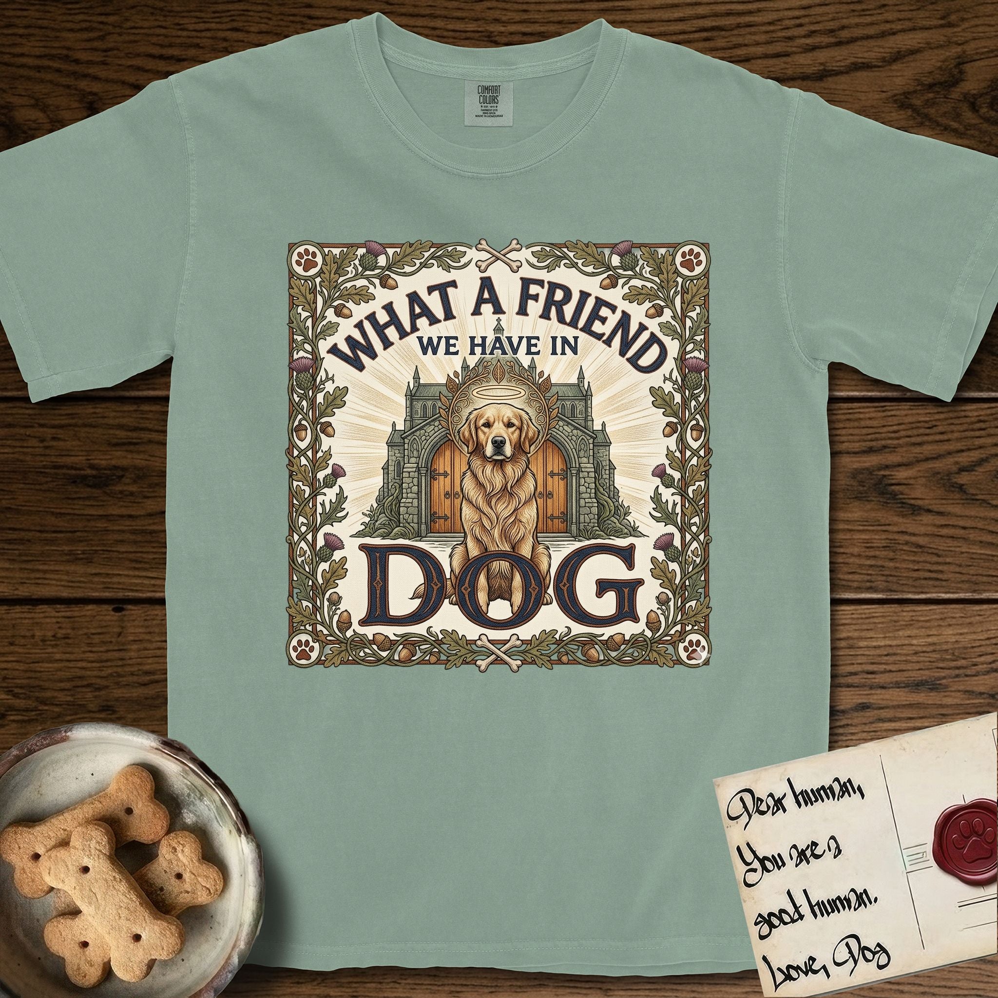 What a Friend We Have in Dog T-Shirt flat lay in Bay, featuring an Arts and Crafts tapestry illustration of a haloed Golden Retriever seated before a monastery, framed by an oak-leaf, acorn, and thistle border, with the word DOG below.